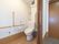 洋式のトイレが備わる空間は、白色の壁、淡い茶色のフローリングの床である。手すりや緊急時呼び出しブザーが設けられている。