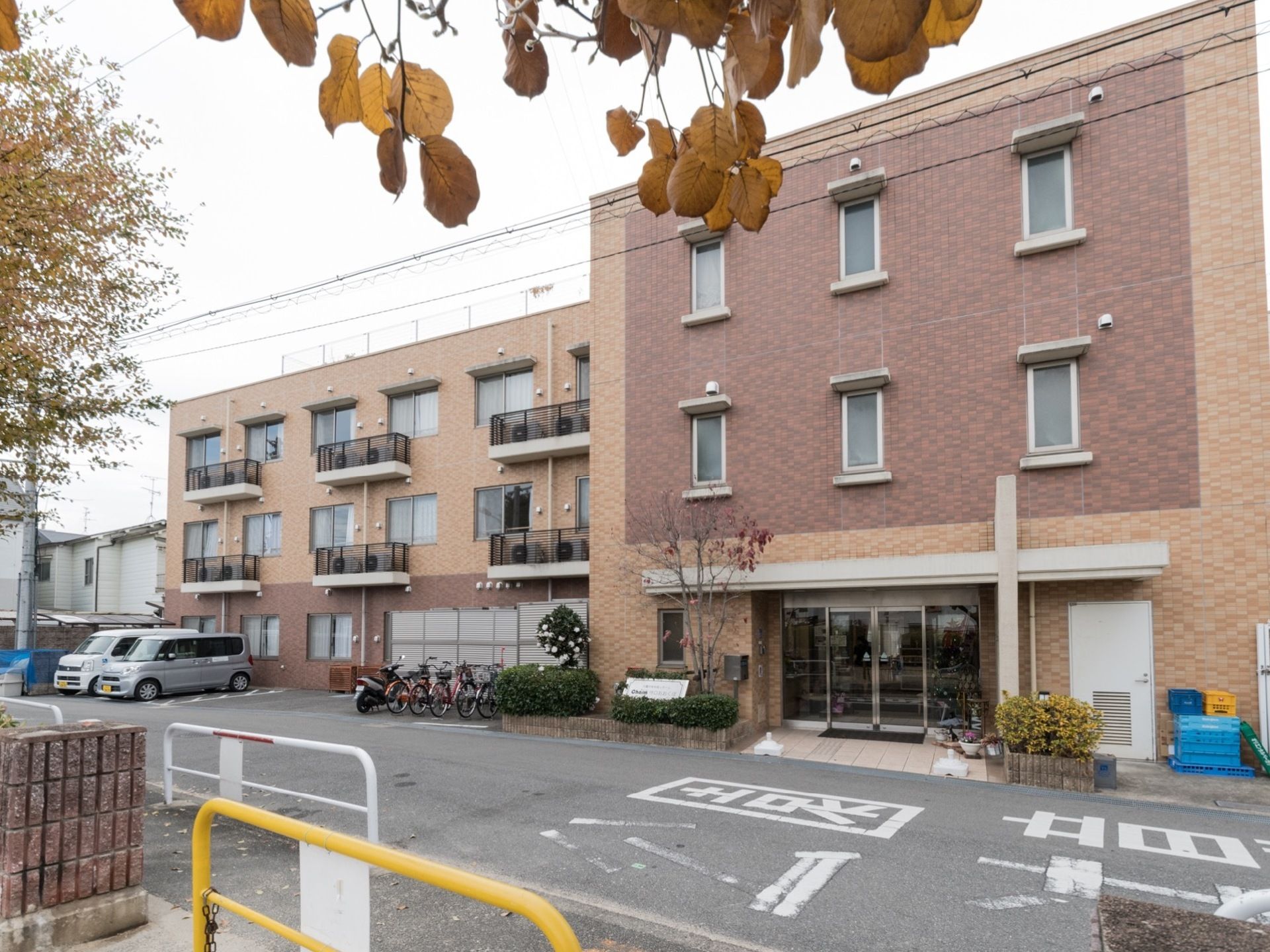 落ち着いた色合いの外壁の施設は３階建てである。施設前に駐輪スペースや駐車場があり、来訪時に利用できる。