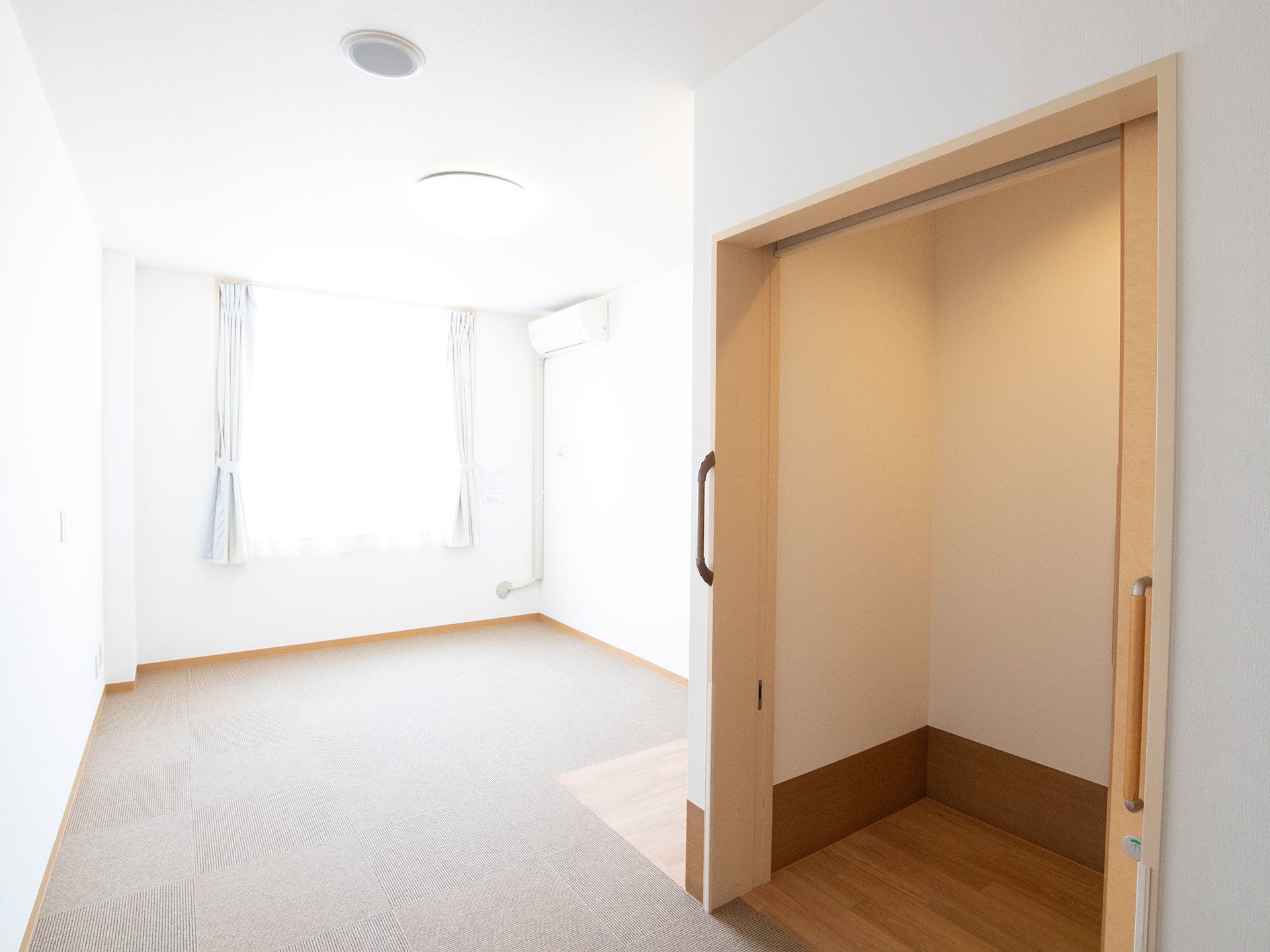 居室の壁、窓につけられたカーテンは白で統一されている。床にはグレーのタイルカーペットが敷かれている。