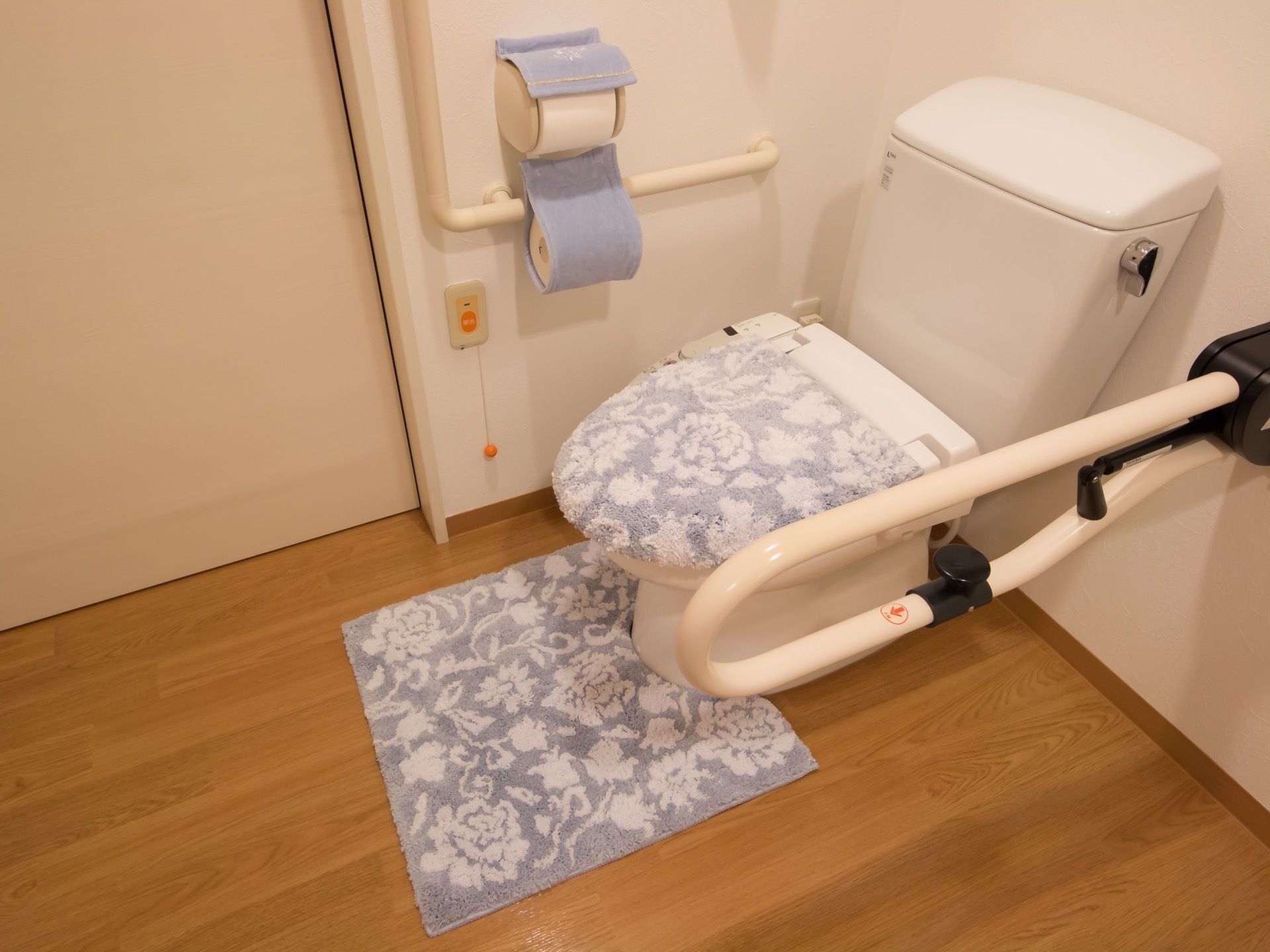 トイレマットと、便器フタカバーのついた洋式トイレである。便座の両サイドには、手すりがついており、壁には呼出ボタンがある。
