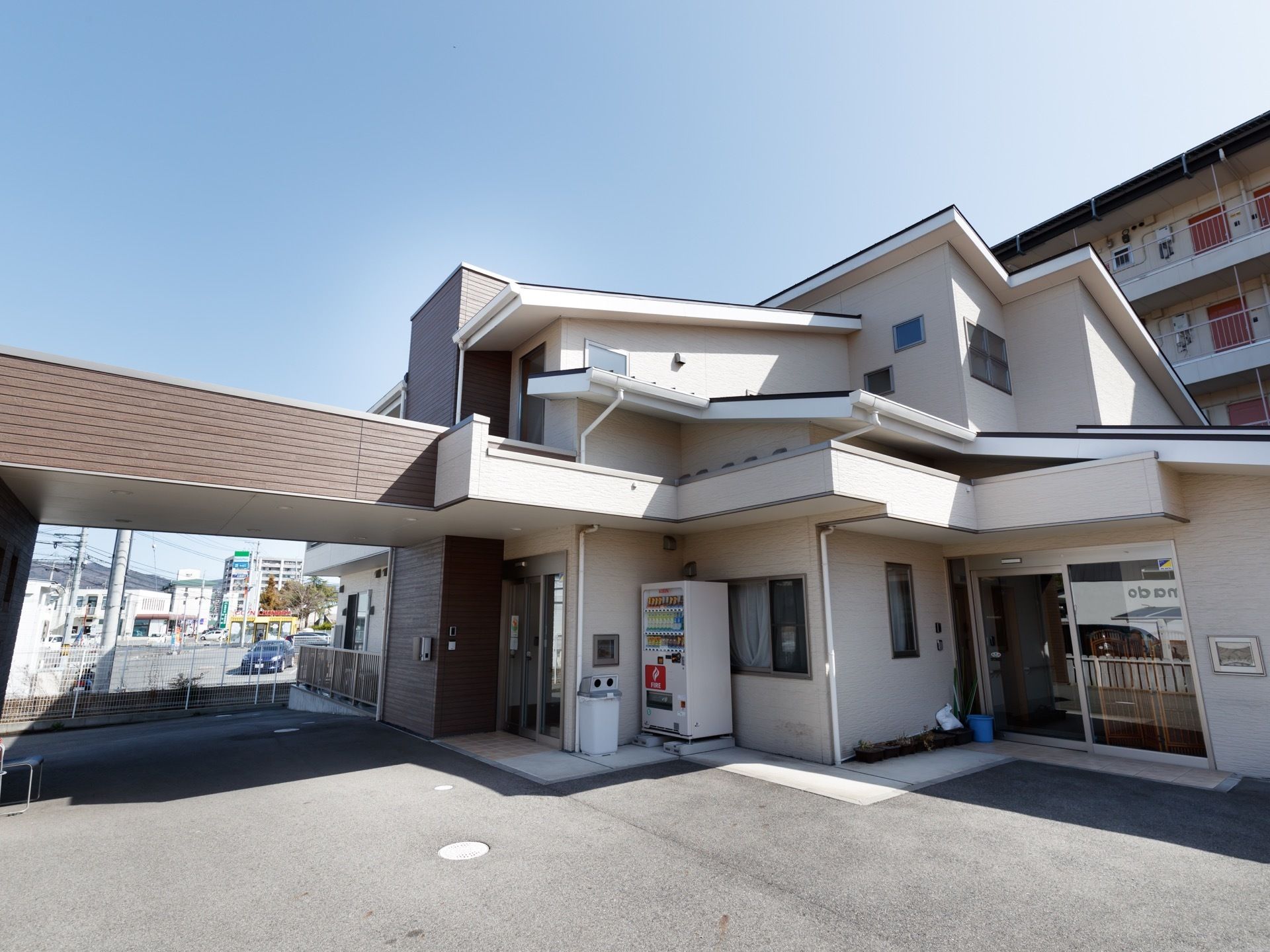 玄関前は広くバリアフリーになっている。扉の近くには自動販売機が設置されている。施設の後ろに階数の低いマンションがある。