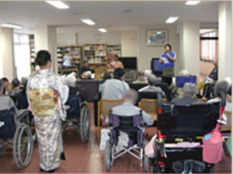 椅子や車いすに座った20名ほどのご老人が多目的ホールに集まりスタッフの話を聴いている風景
