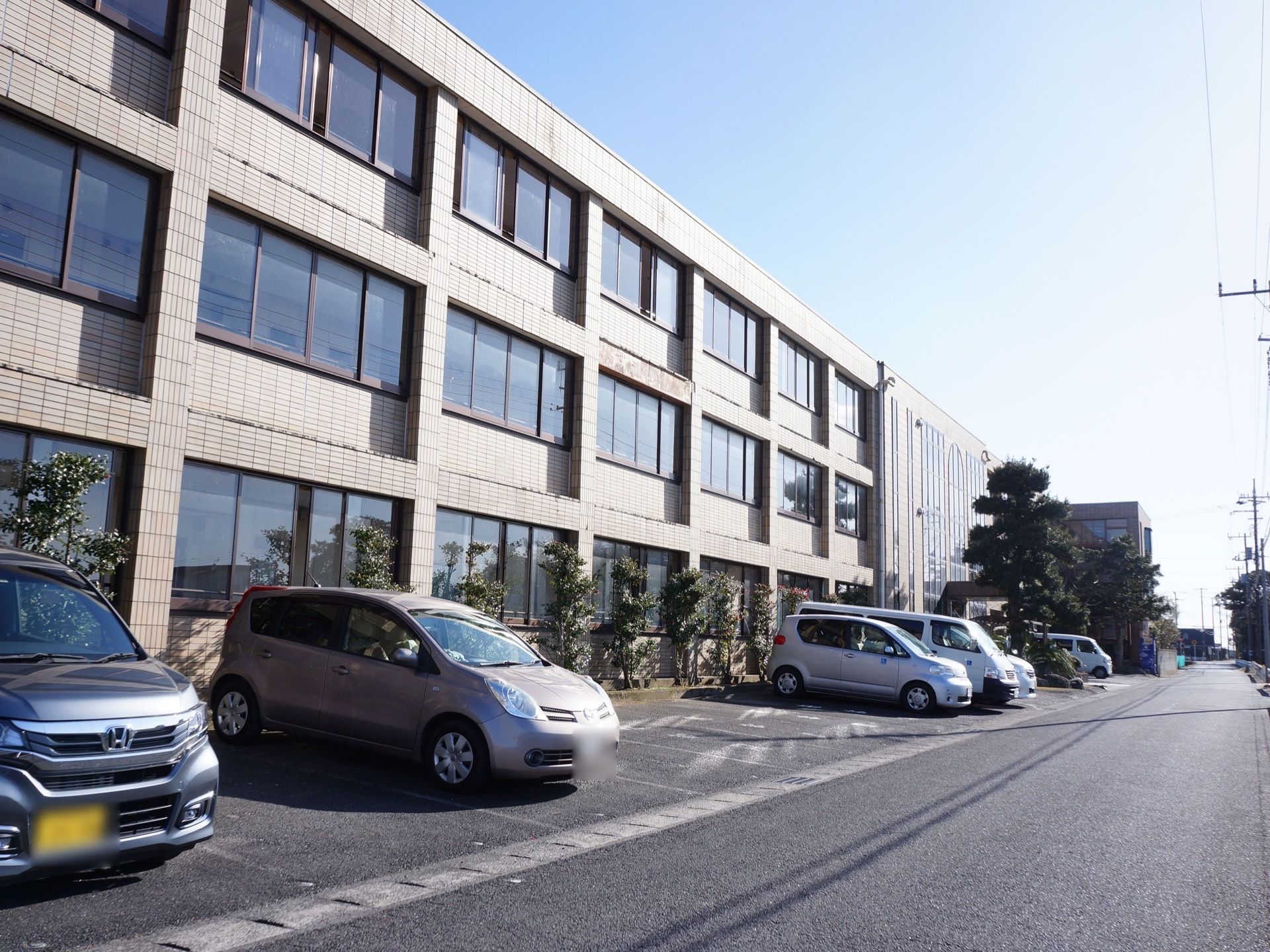 施設は3階建ての建物。道路と建物の間は白線で区切られた駐車スペースになっている。複数の車が駐車している。