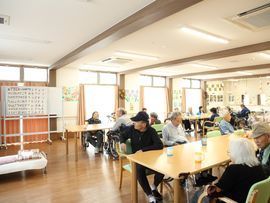 洗面台が並んだ食堂には入居者が集まっている。ホワイトボードに貼られた紙には「ありさんあつまれ」から始まる言葉が書かれている。