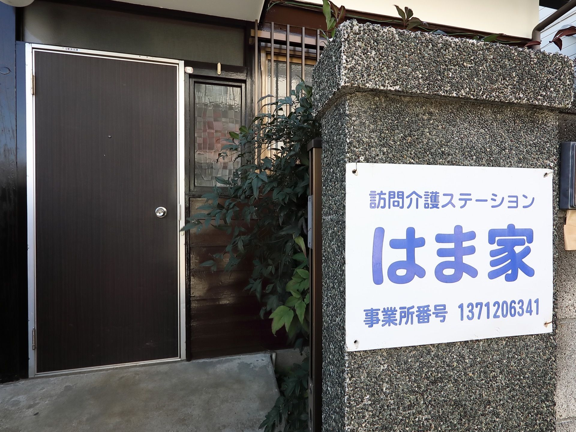 片開きのドアで、1軒屋のような外観の建物である。さまざまな植物が植えられている。玄関前の石柱にプレートが貼られている。