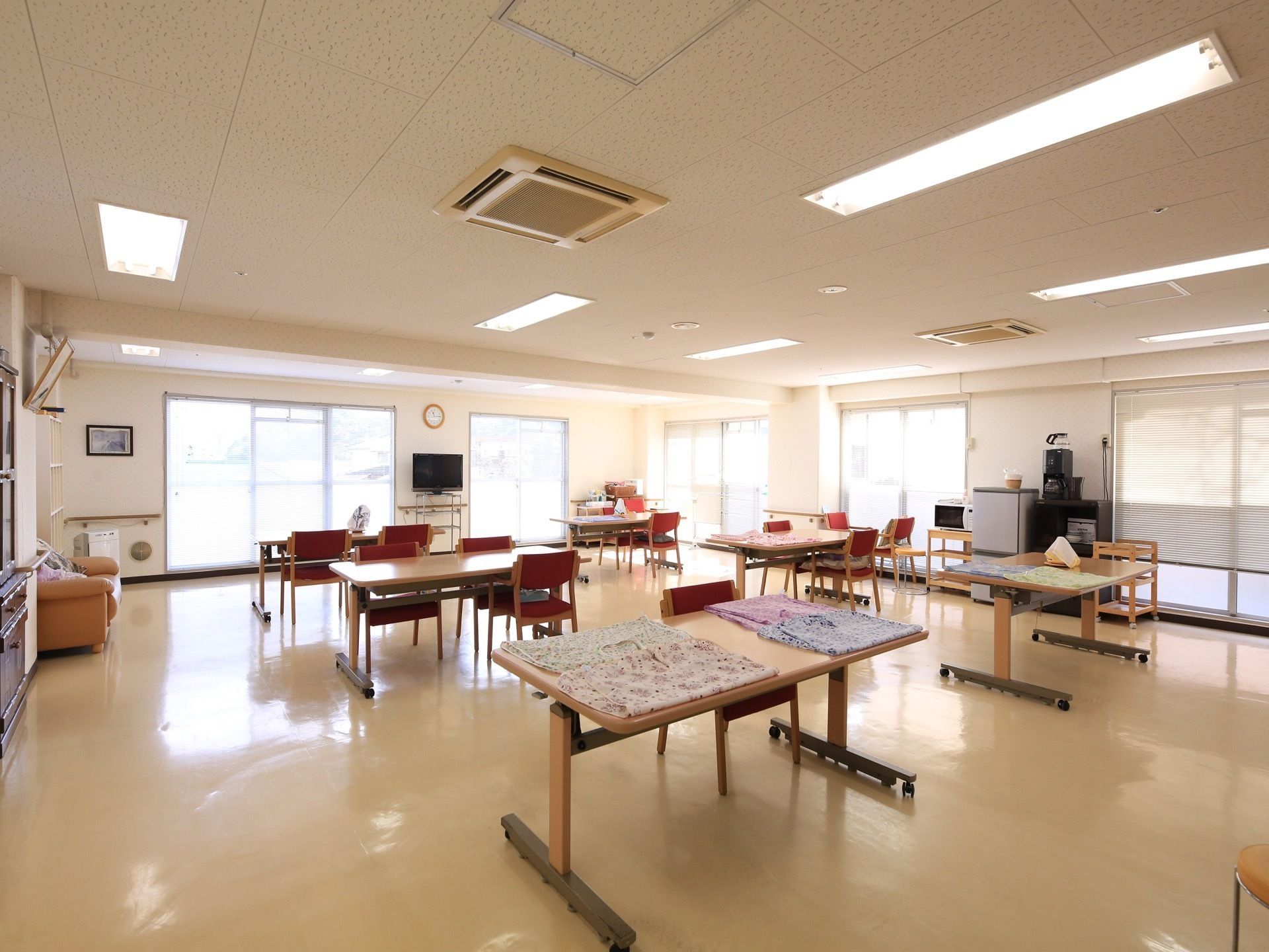 広々とした共有スペースで、Ｌ字型にたくさんの窓が開いている。壁際にオレンジのソファがある。奥に冷蔵庫やレンジが置かれている。