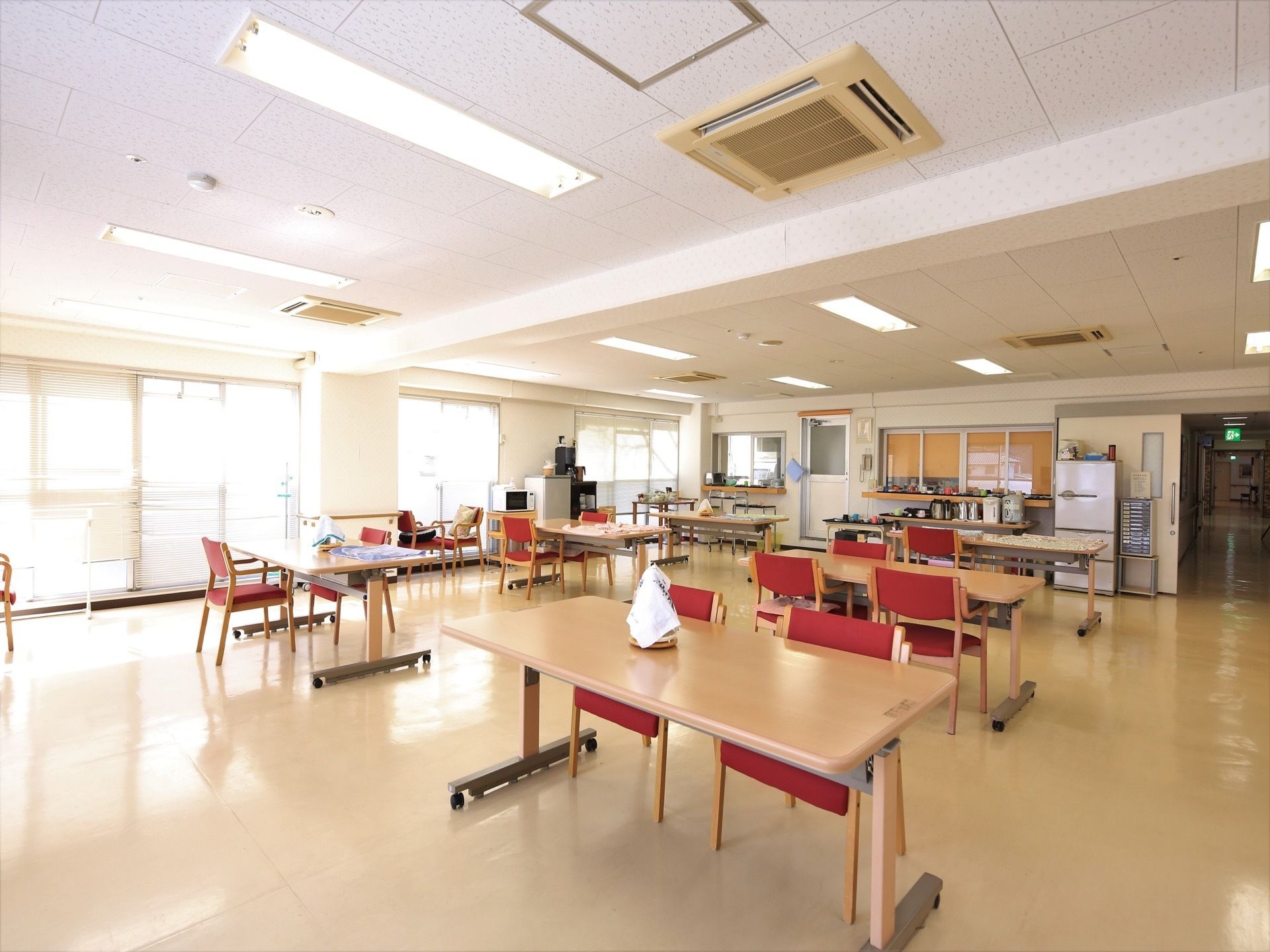 窓がたくさん並び、とても明るいダイニングである。赤い椅子のテーブルセットがある。壁際に冷蔵庫が置かれている。