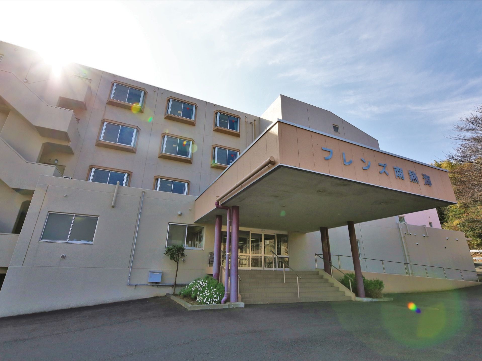 施設名が書かれた屋根が設置されている玄関で、両サイドに花壇がつくられている。目の前の道はアスファルトで舗装されている。