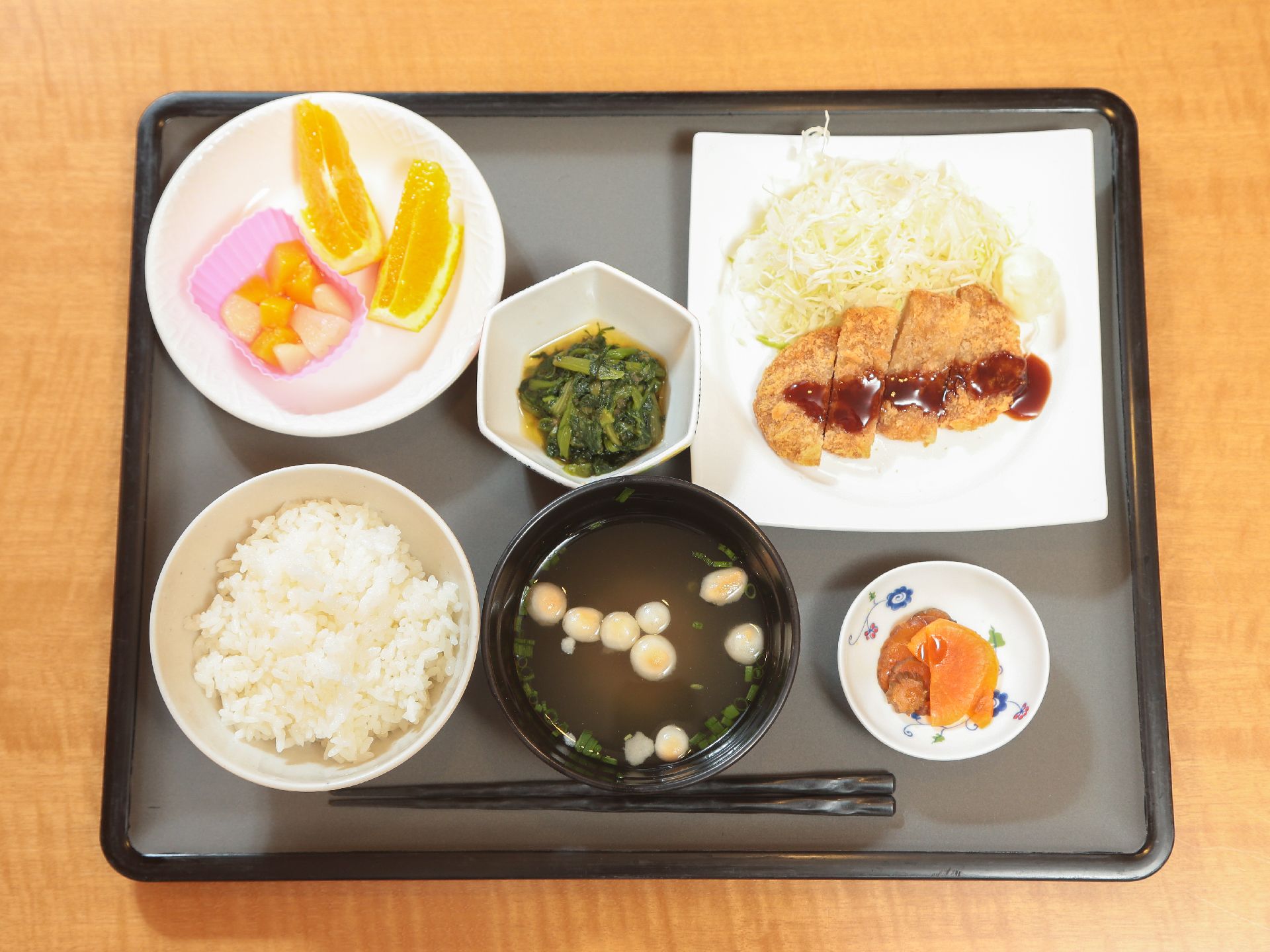 グレイのトレーにご飯と汁物、メインのおかずに小鉢1つ、デザートのフルーツとお漬物が添えられている。1食の献立である。