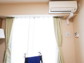 緑色のカーテンがかかる窓からは陽光が差し込んでいる。エアコンが右上に取り付けられており、左手にはランプのシェードがみえる。