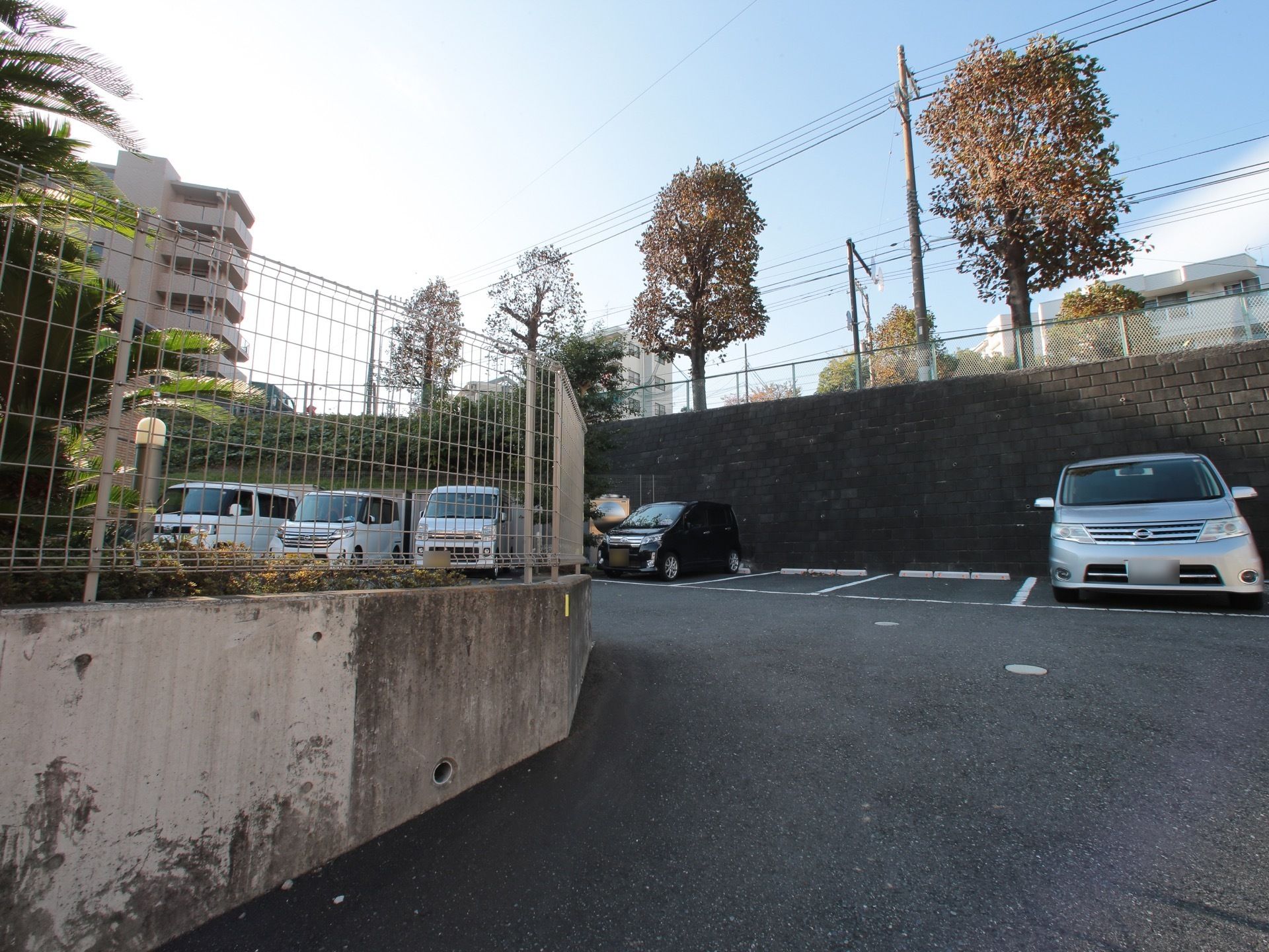 駐車場の入り口は傾斜がついており、周囲にはフェンスが設置されている。奥には、複数台の車が駐車されている。