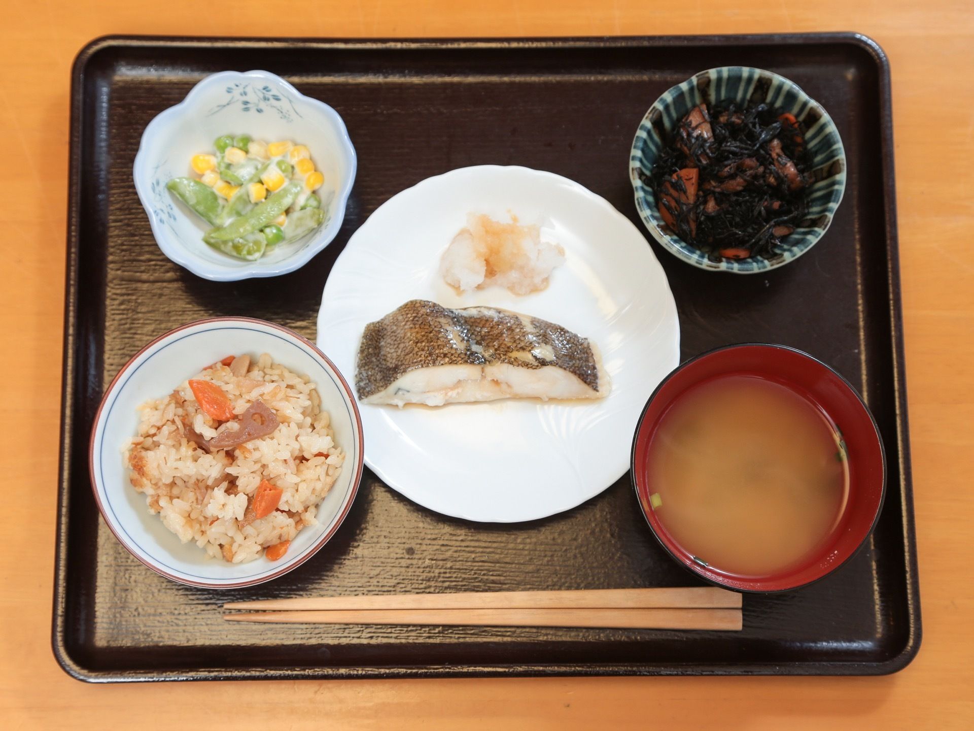 テーブルの上に置かれた黒いトレイにみそ汁や炊き込みご飯、主菜や２種類の副菜が並べられている。箸も用意されている。