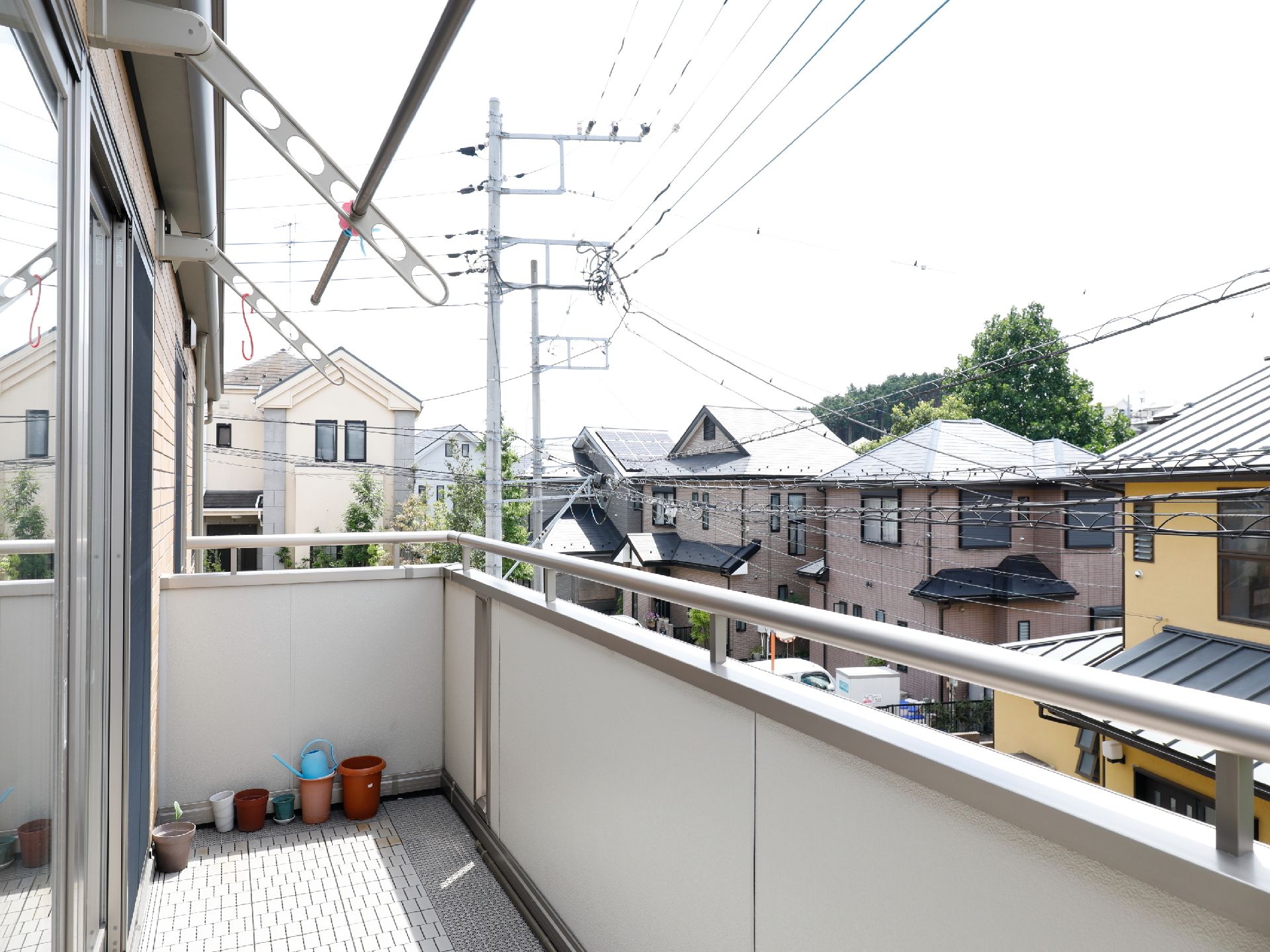 部屋から外に出たベランダスペースで、タイル調の床で白色のフェンスに上部はシルバーの手すりが付いている。物干し竿があり、植木鉢が置いてある。
