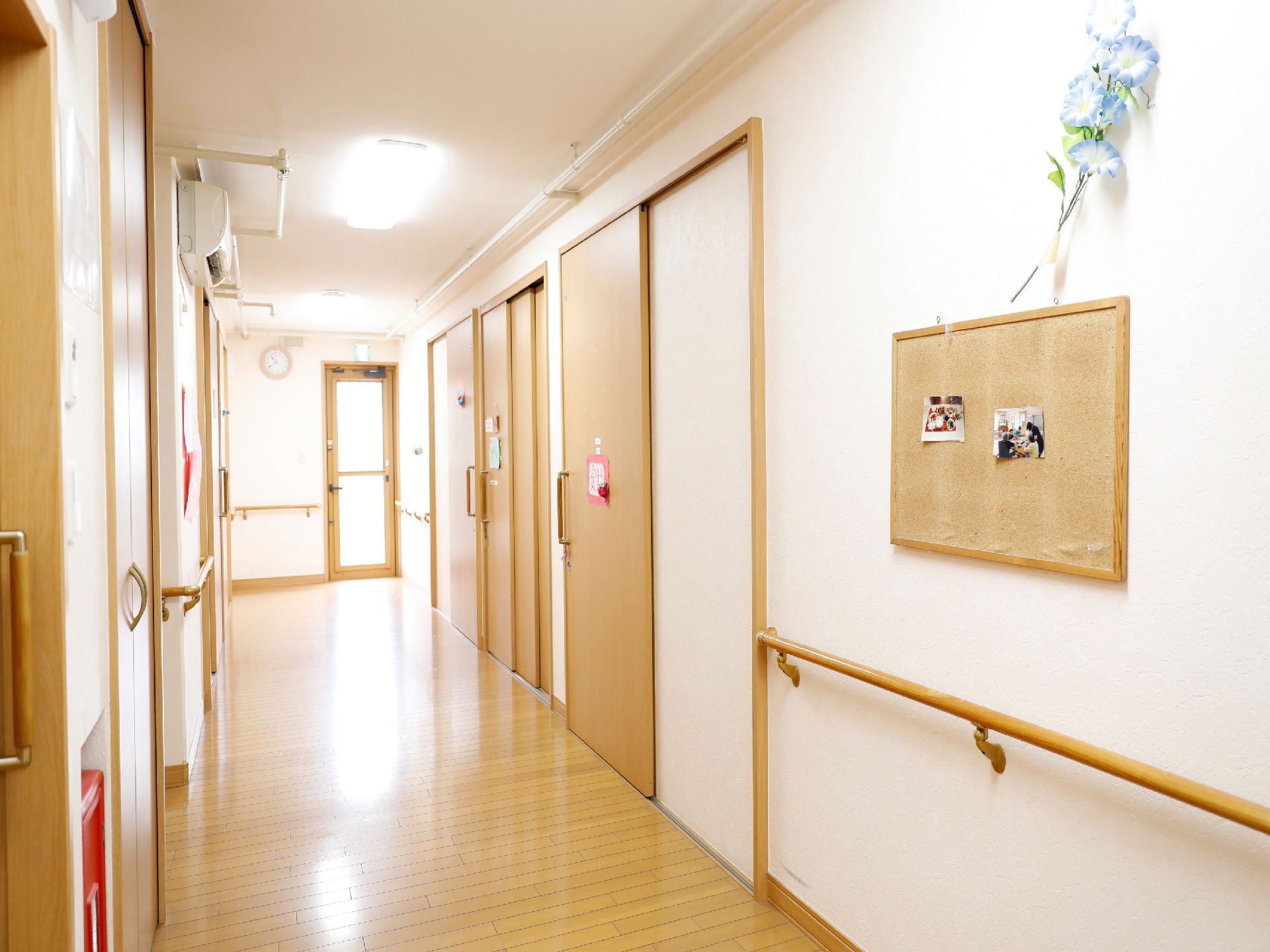 白色と木目仕様の通路、壁には手すりが設置してあり、天井下にはエアコンが取り付けてある。壁には、写真を貼ったボードや花が飾られている。