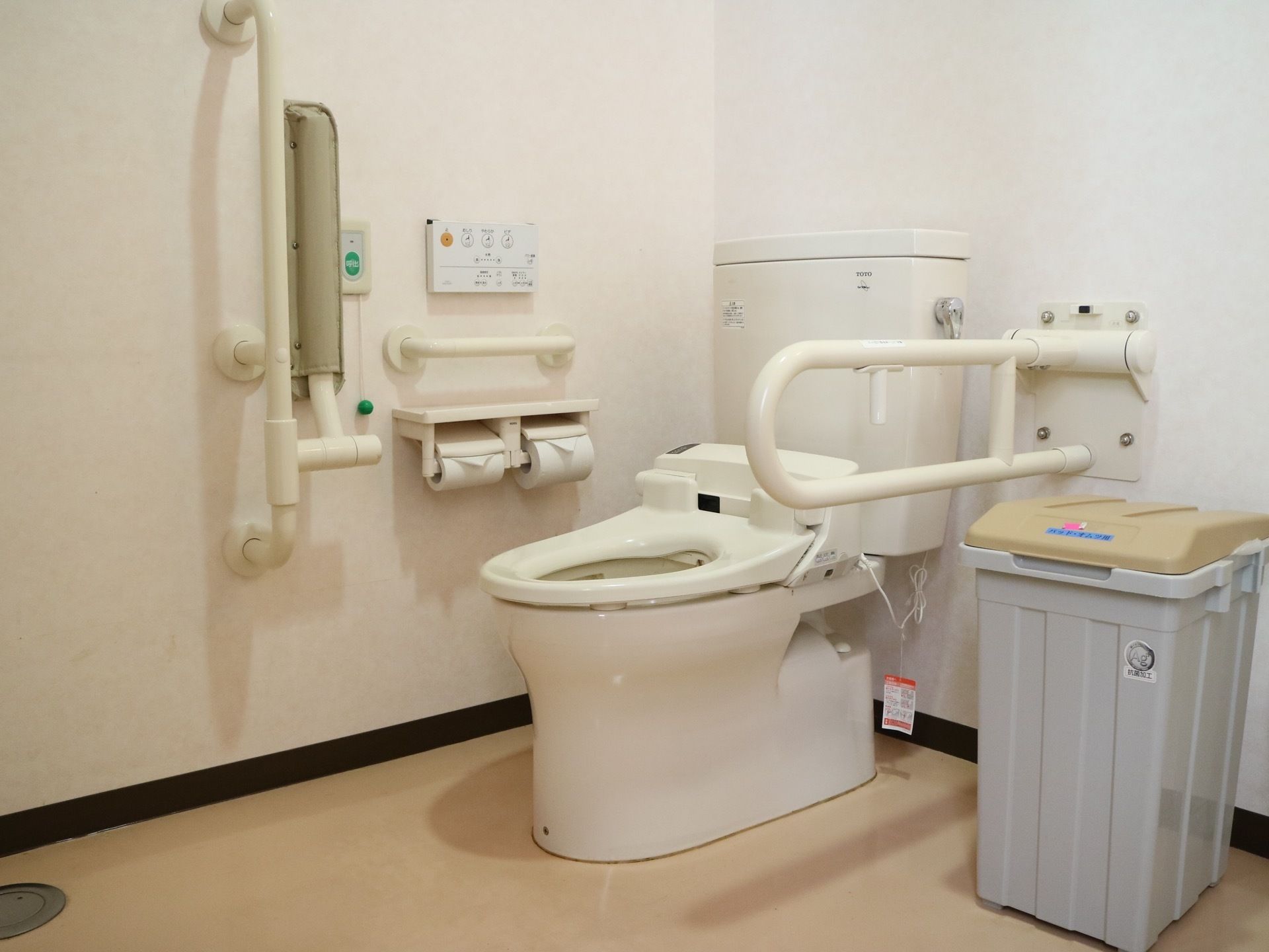 蓋が付いていない洋式便器が設置されている。便器の両側にさまざまな形状の手すりが用意されており、壁面には呼び出しボタンがある。