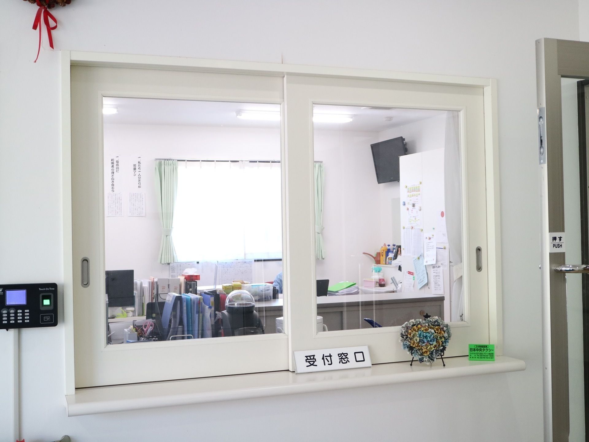 ガラス戸が設置された受付カウンターがあり、奧に事務室の様子が見える。窓の手前に、タクシー会社の電話番号案内と手作りのオブジェがある。