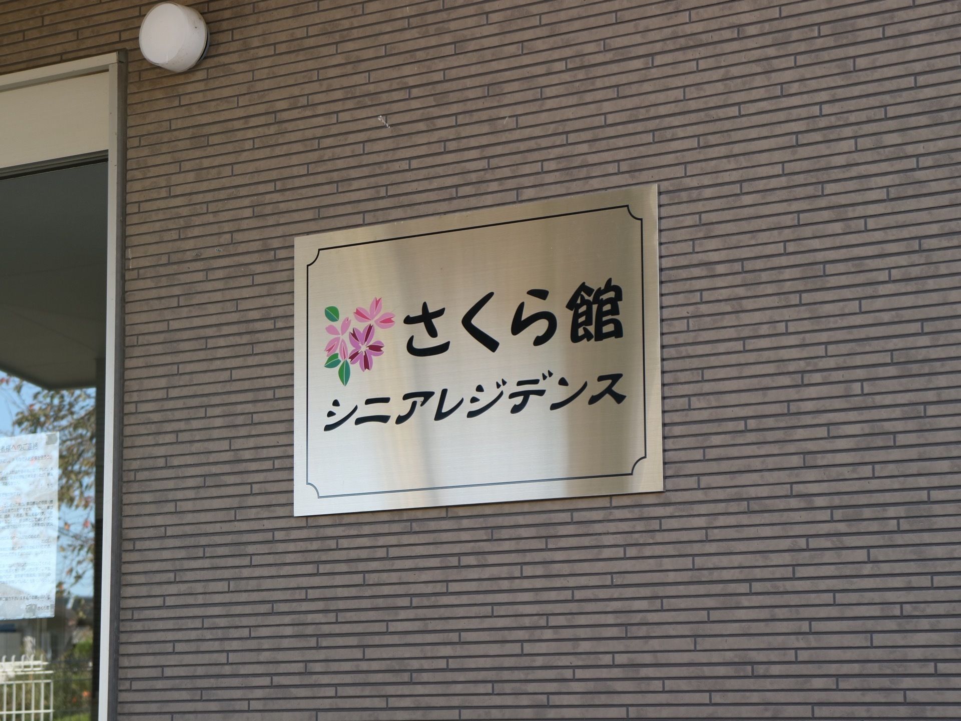 外壁に取り付けられている四角い金属プレートである。施設名称が明記されており、桜のイラストが描かれている。