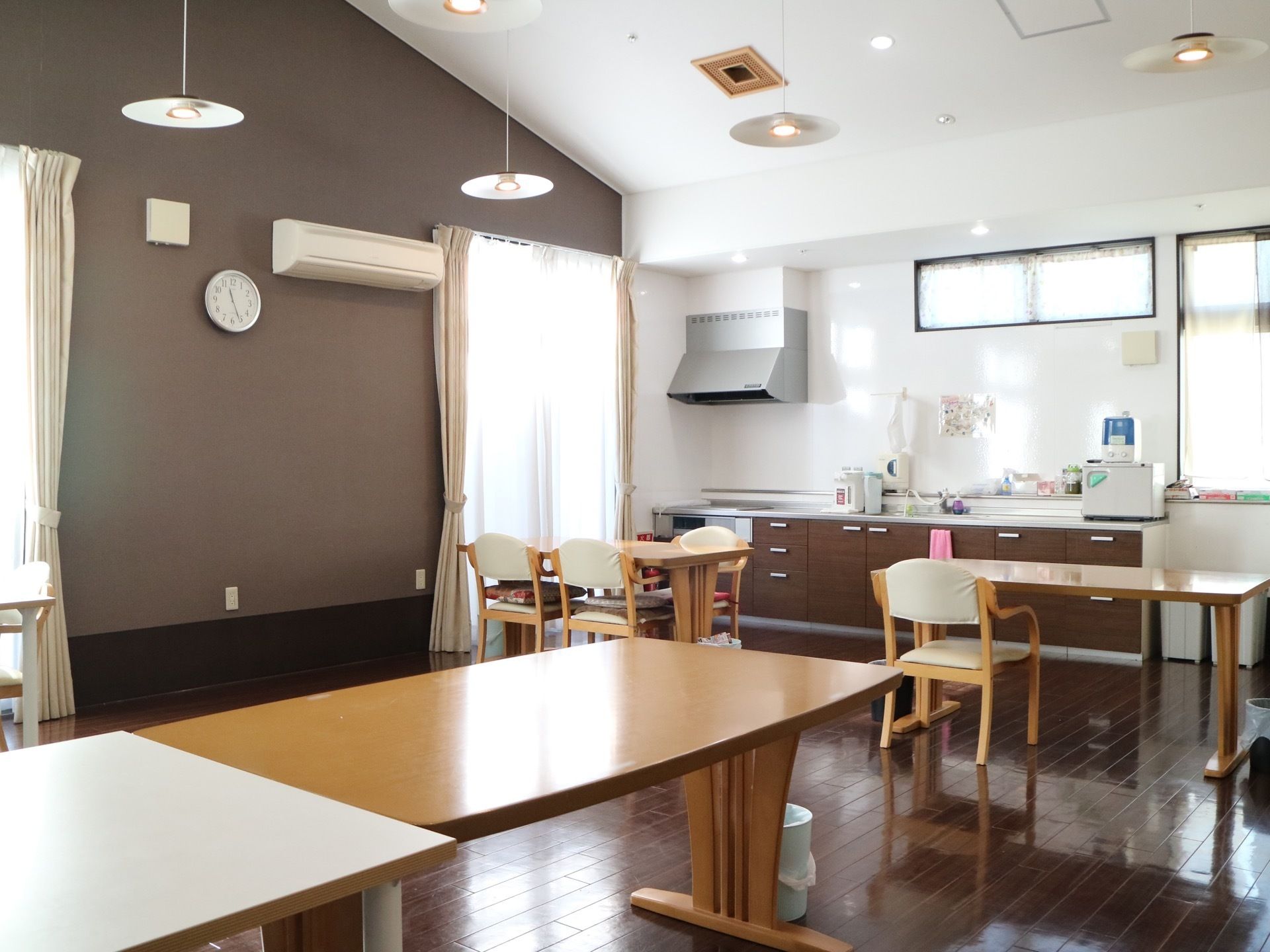 茶色い壁紙が施されている広々とした食堂。テーブルや椅子が設置されている。システムキッチンの上に電気ポットが置いてある。
