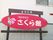 窓が付いている外壁の前に、看板が設置されている。木製の柱が組み合わされている台に、楕円形のパネルが取り付けてある。