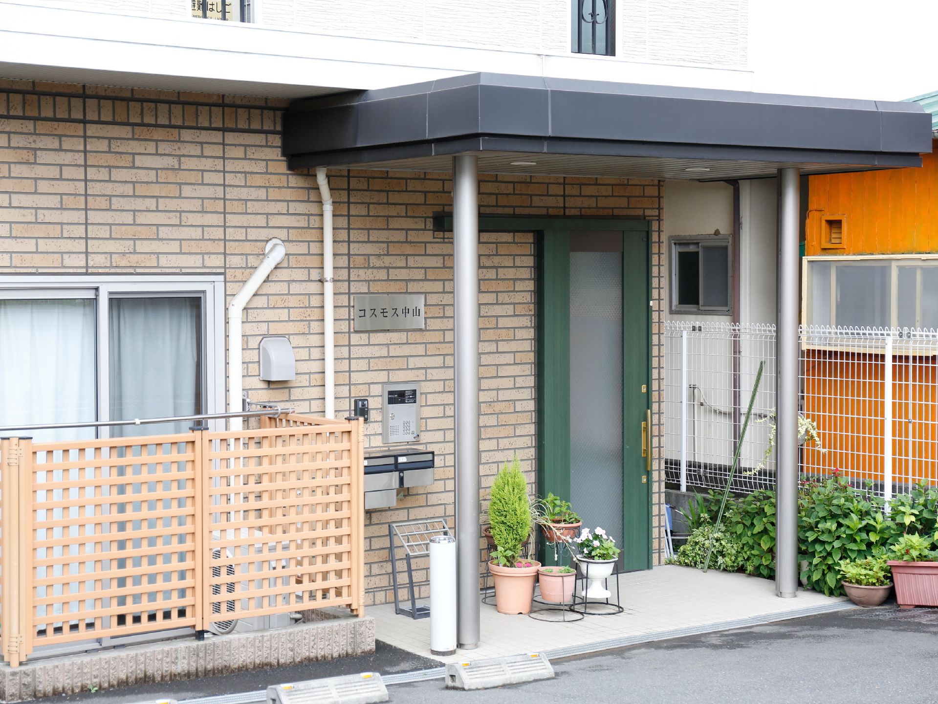 施設の玄関前の床はグレー系のタイル敷きである。花などが植えられた鉢が並び、その左側には傘立てと灰皿が置かれている。