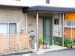 施設の玄関前の床はグレー系のタイル敷きである。花などが植えられた鉢が並び、その左側には傘立てと灰皿が置かれている。
