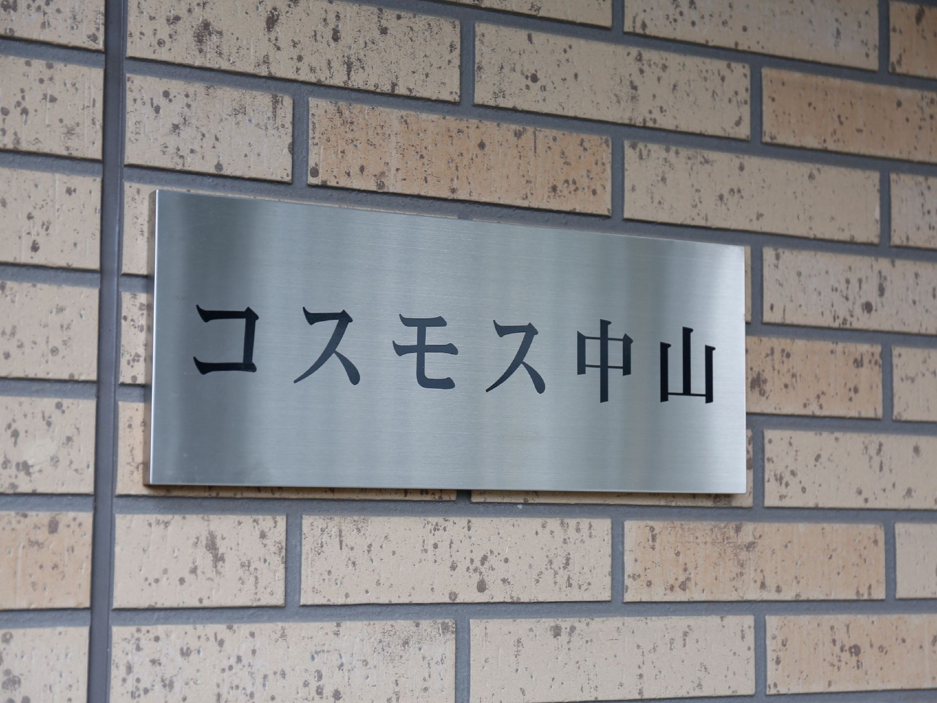 施設の玄関付近の外壁はベージュ系のタイル張りである。その上に施設名が表示された横長のプレートが設置されている。