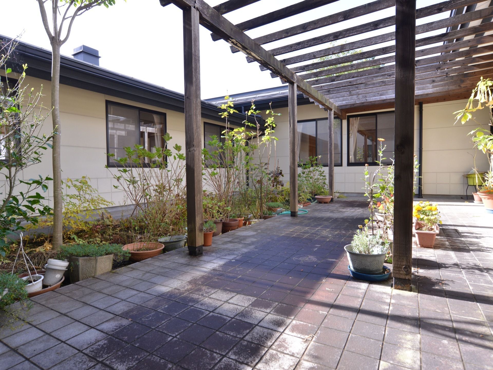 タイル張りの中庭。木製の大きなひさしがかかっている。さまざまな種類の植木が植えられ、鉢植えやプランターには季節の花が咲いている。