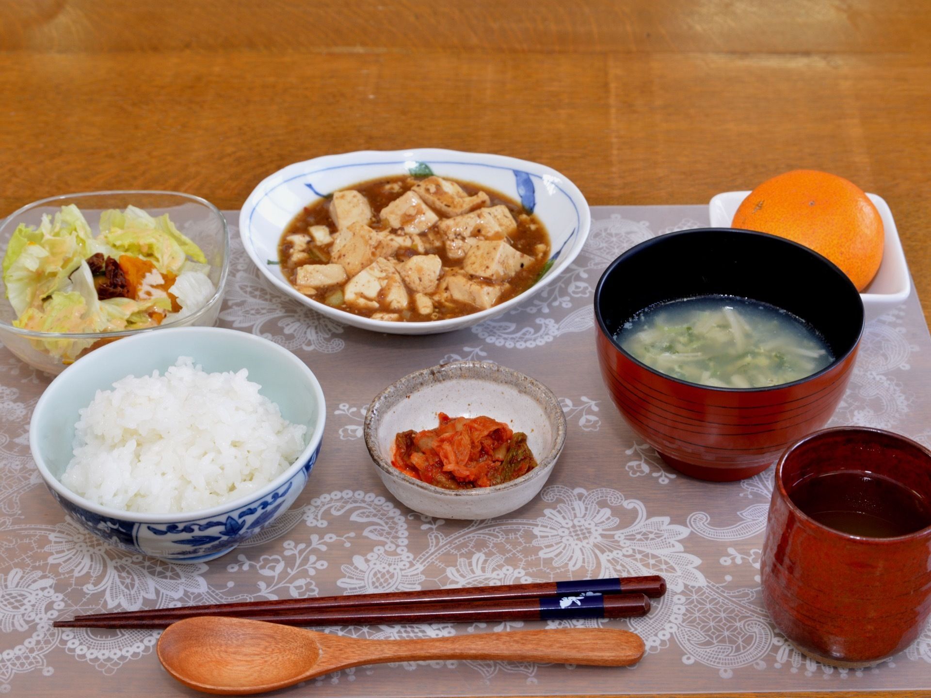 白いご飯、汁物、主菜、副菜、デザートなどが配膳されている。家庭的なメニューで、スプーンが添えられている。