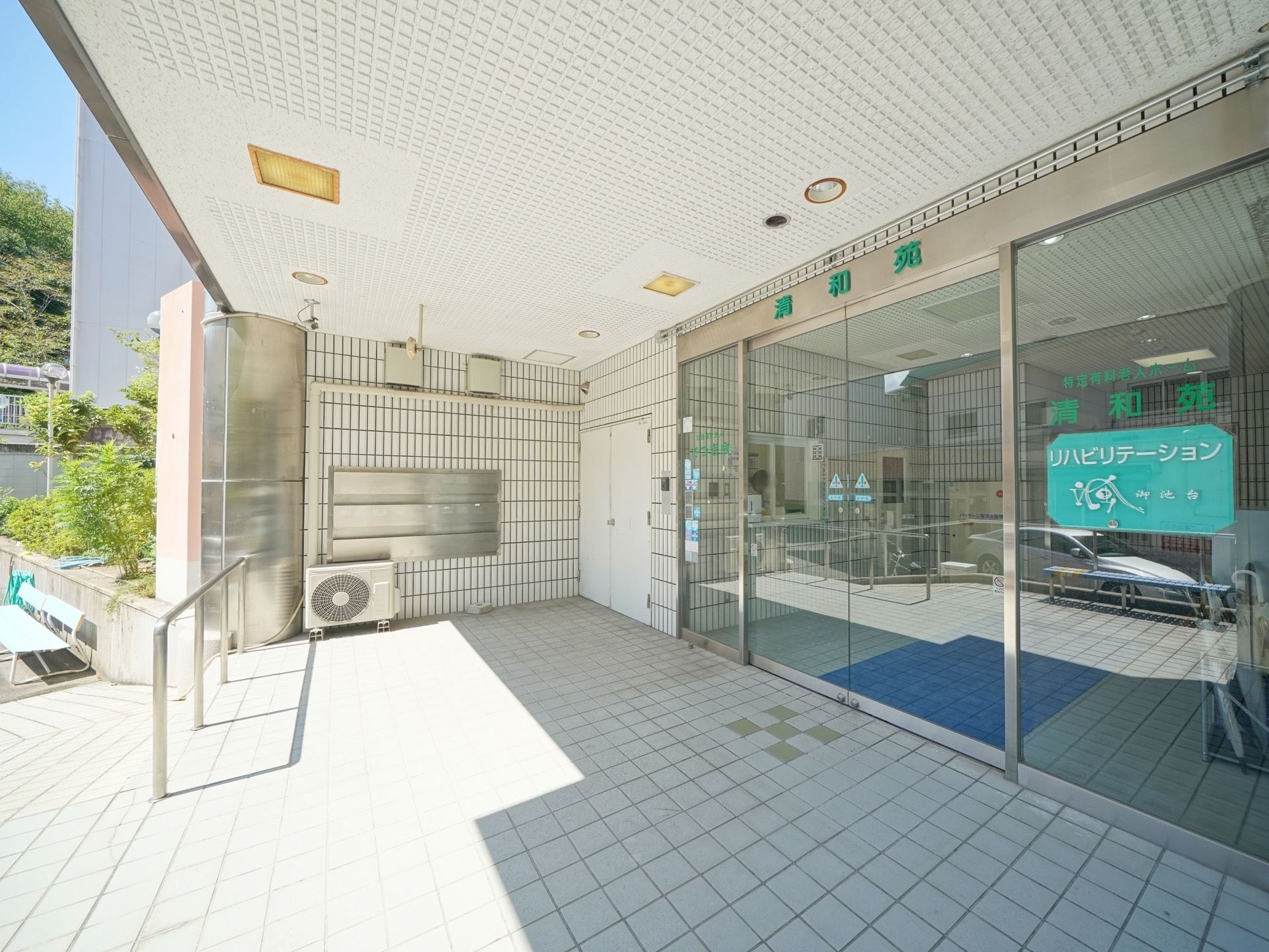 大きな自動扉の玄関である。玄関には施設名がはっきりと掲示されている。駐車場からのスロープ脇には手すりが設置されている。