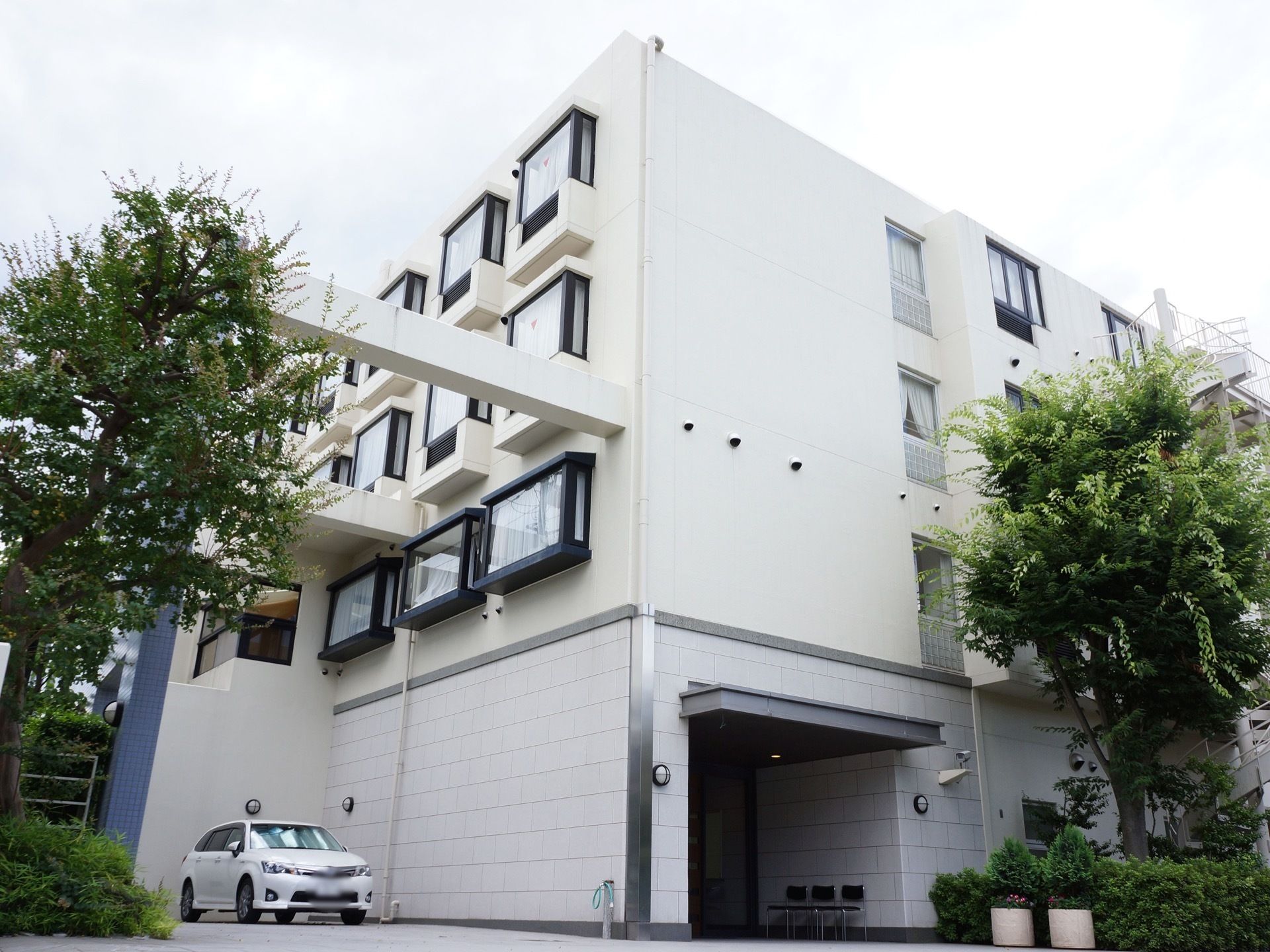 植木や低木に囲まれ、黒い出窓が外壁に並ぶホワイトの建物