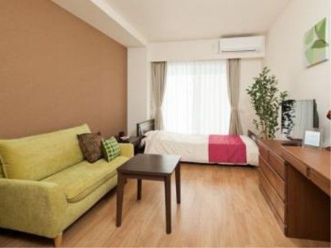 窓際にベッド、黄緑の長ソファーとブラウンのチェストとデスクが置かれた部屋