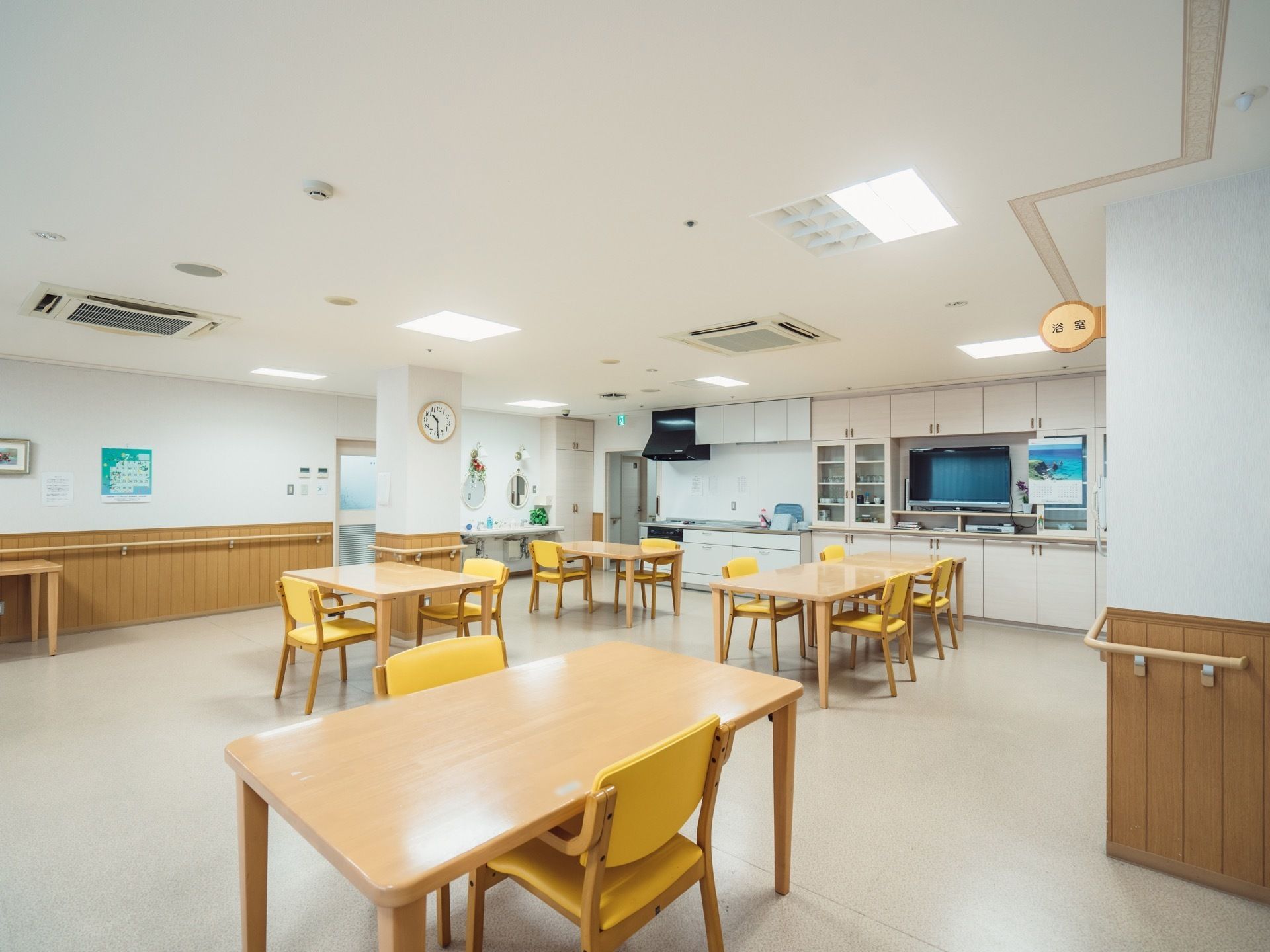 ダイニングテーブルがたくさん用意されている食堂である。部屋の奥にはキッチンが併設されている。壁には手すりが備わっている。
