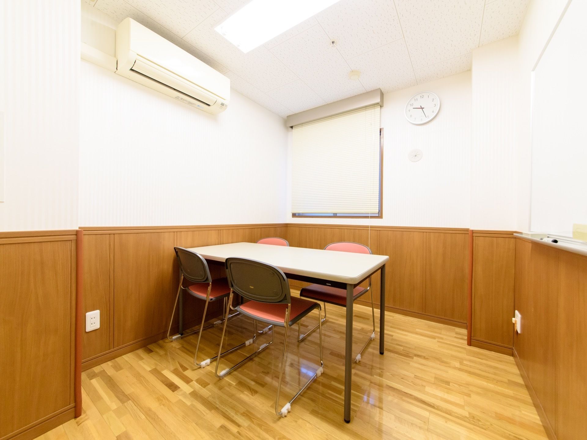 談話室の窓にはブラインドカーテン、壁には時計がかけられている。白いテーブルの周りにはピンク色の背もたれ付きの椅子が並べられている。