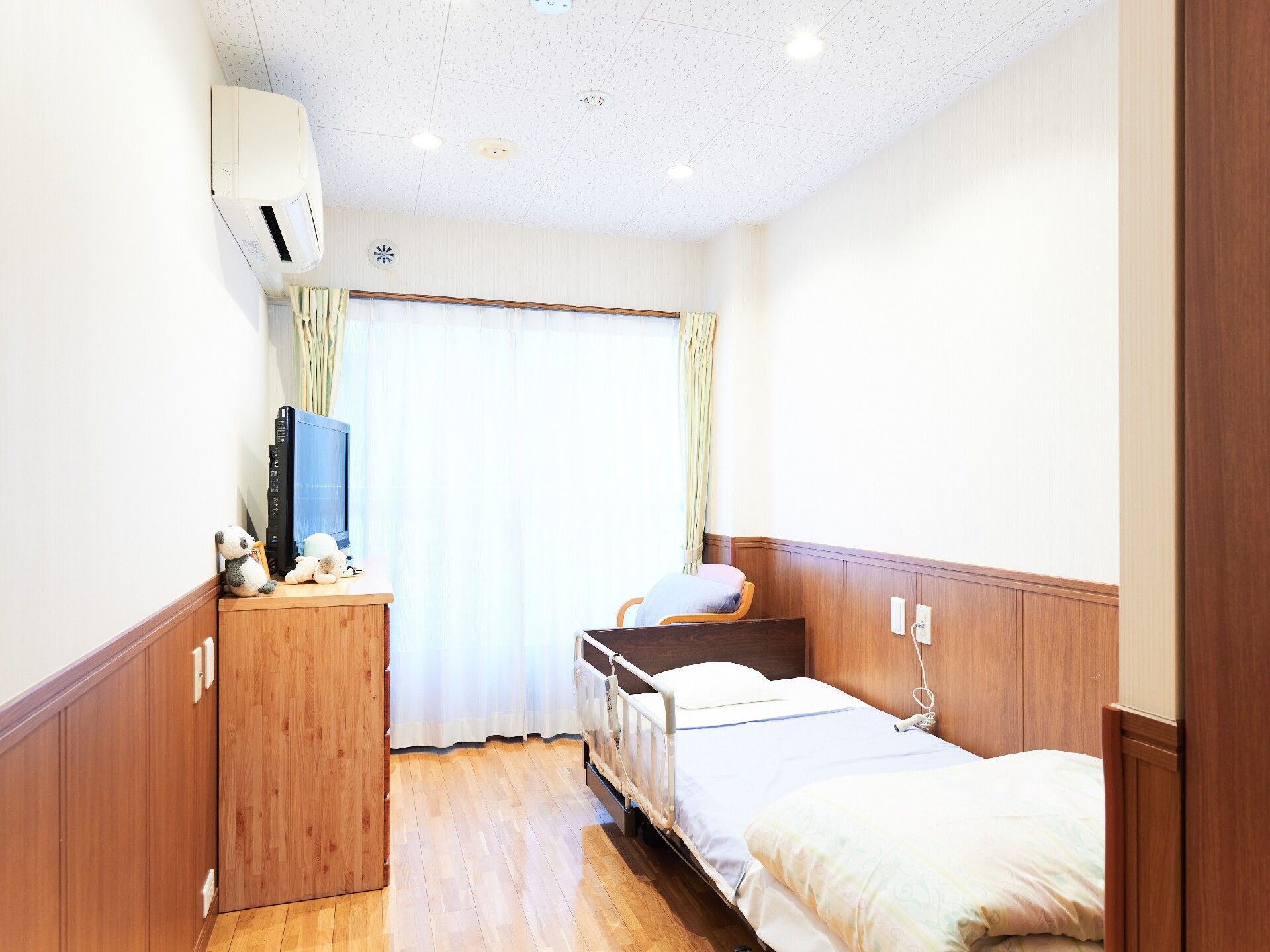 居室には介護用ベッドが１台置かれている。ベッドには柵が付いており、横の壁にはコンセントとスイッチがある。
