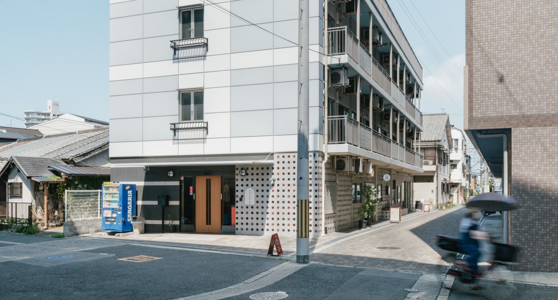 施設は４階建てである。１階には自動販売機が設置されている。その近くと建物の側面側には車が停められている。