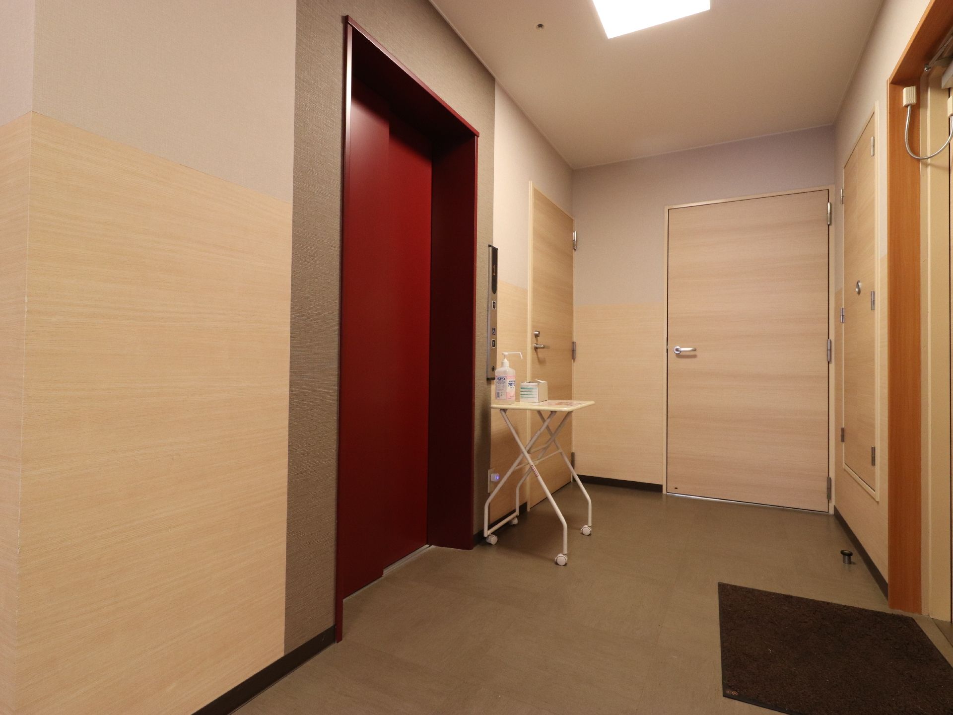 エレベーターの前に、手指に使用するアルコール等の消毒薬が置かれている。また、エレベーターの扉は落ち着いた赤色で雰囲気になじんでいる。