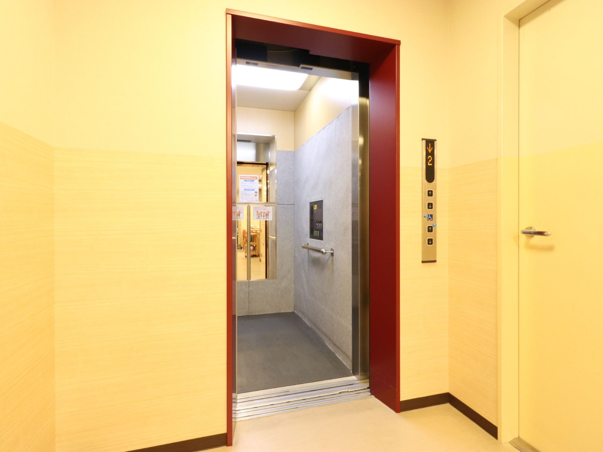 エレベーターがあり、周辺の壁は黄色、エレベーターの淵は赤い色になっている。扉が空いていて、内部が見える。