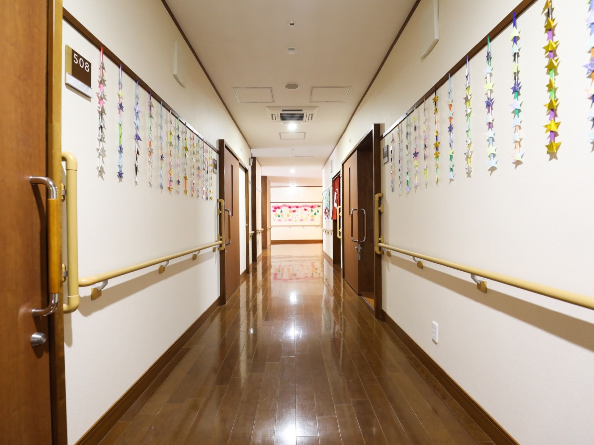 施設の廊下はフローリング敷きで、ダークな色合いで光沢のある素材が使われている。壁の両側には手すりが設けられている。