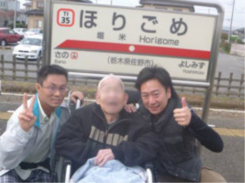 駅のホームにある標識前で撮影した職員2人と車椅子の入居者