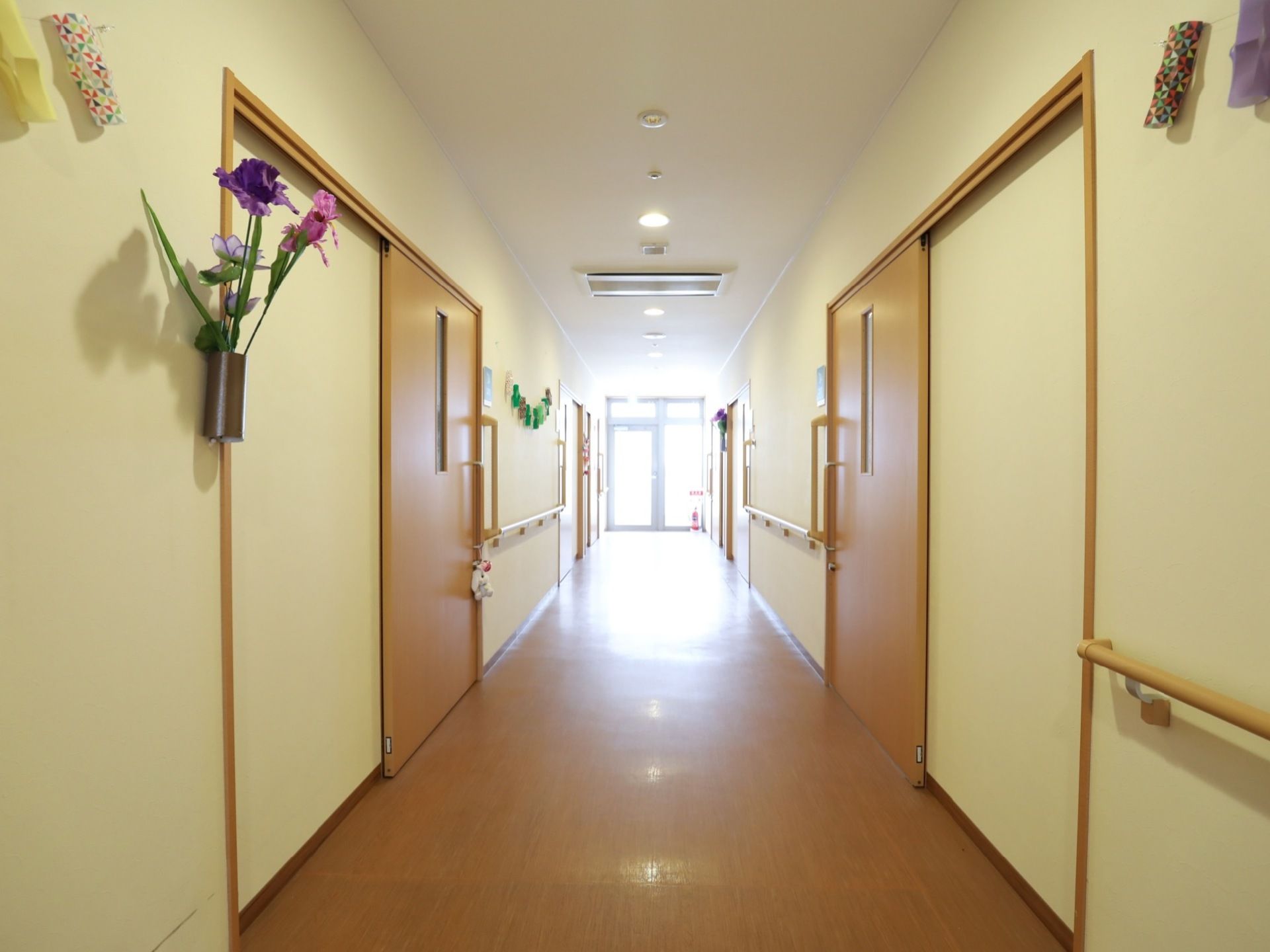 左右に居室ドアの並ぶ施設内の廊下。壁には手すりが設けられ、一番奥の突き当りは非常口のドアになっている。