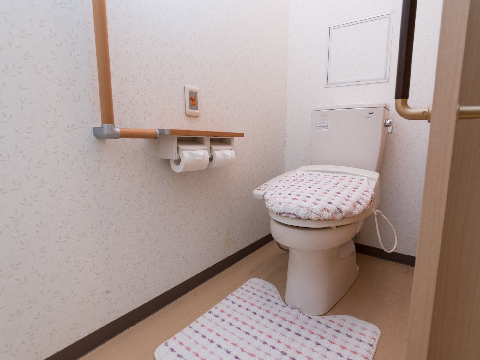 洋式トイレは、便座カバーが取り付けてある。マットも同じ柄で敷いてある。非常用ブザーが壁に取り付けてあるほか、手すりが壁についている。