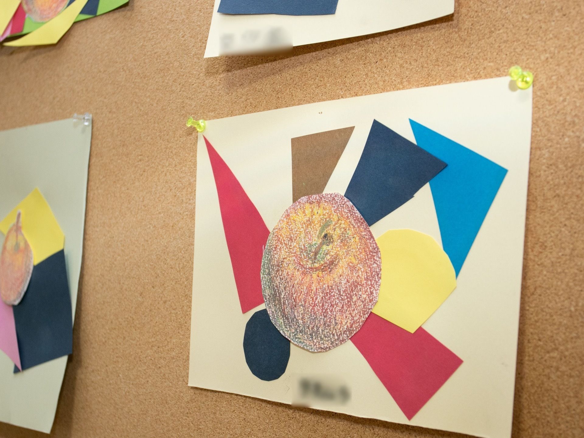 切り紙を使った壁面飾りが飾られている。白い画用紙に、ピンクや青、クリーム色などの自由な形の画用紙を貼り、中央にリンゴの絵が貼られている。