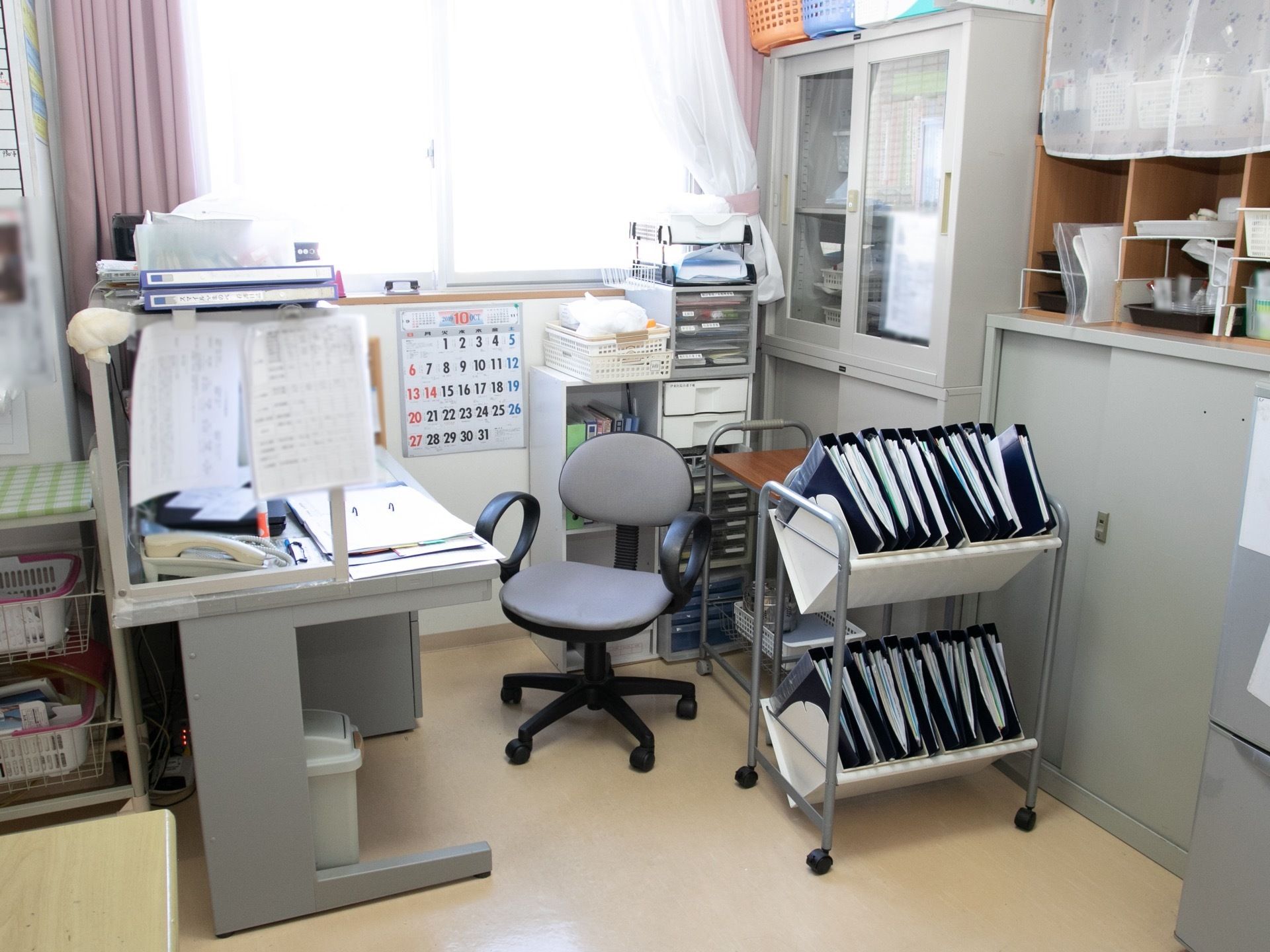 事務室に、事務用デスクと椅子が置かれ、棚やワゴンが並んでいる。ワゴンの中には、資料を挟んだファイルが収まっている。