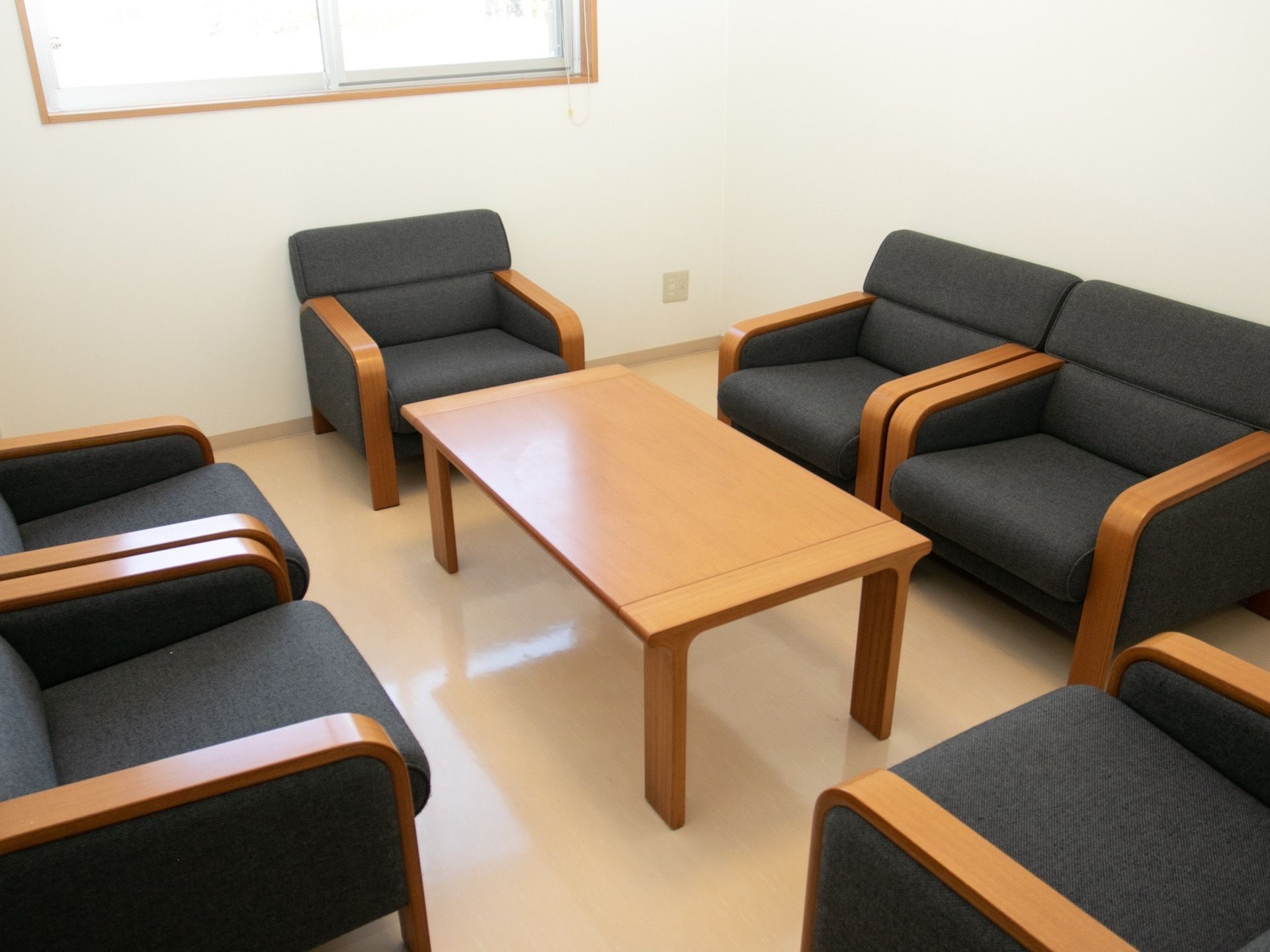 相談室の中央にローテーブルがあり、その周りを囲むように、木製のひじ掛けにグレーの布張りの１人掛けソファが６脚用意されている。