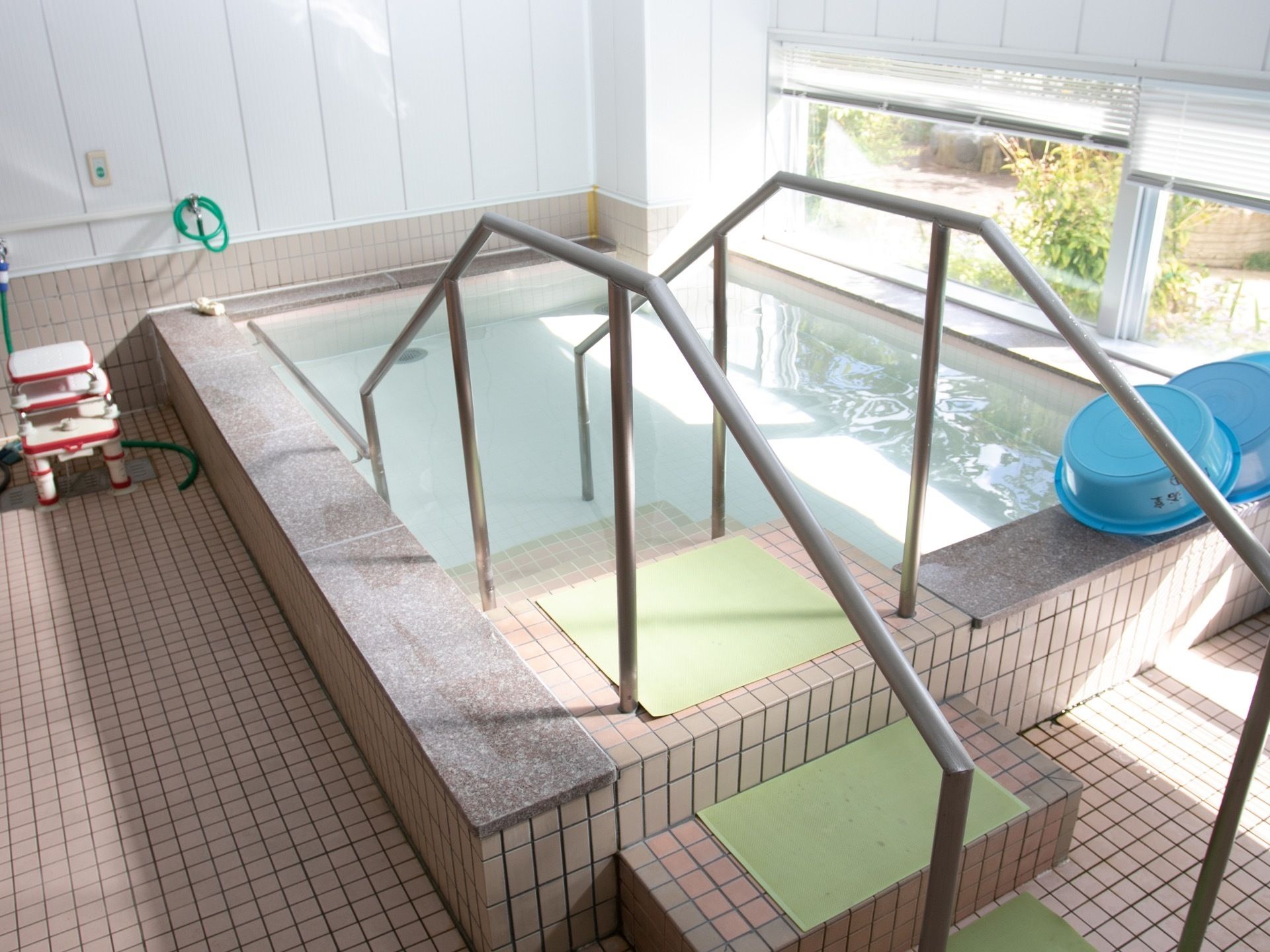 ピンクのタイルが張られた大浴場である。階段と手すりが設置された大きな浴槽があり、お湯が張られている。窓にはブラインドが取り付けられている。