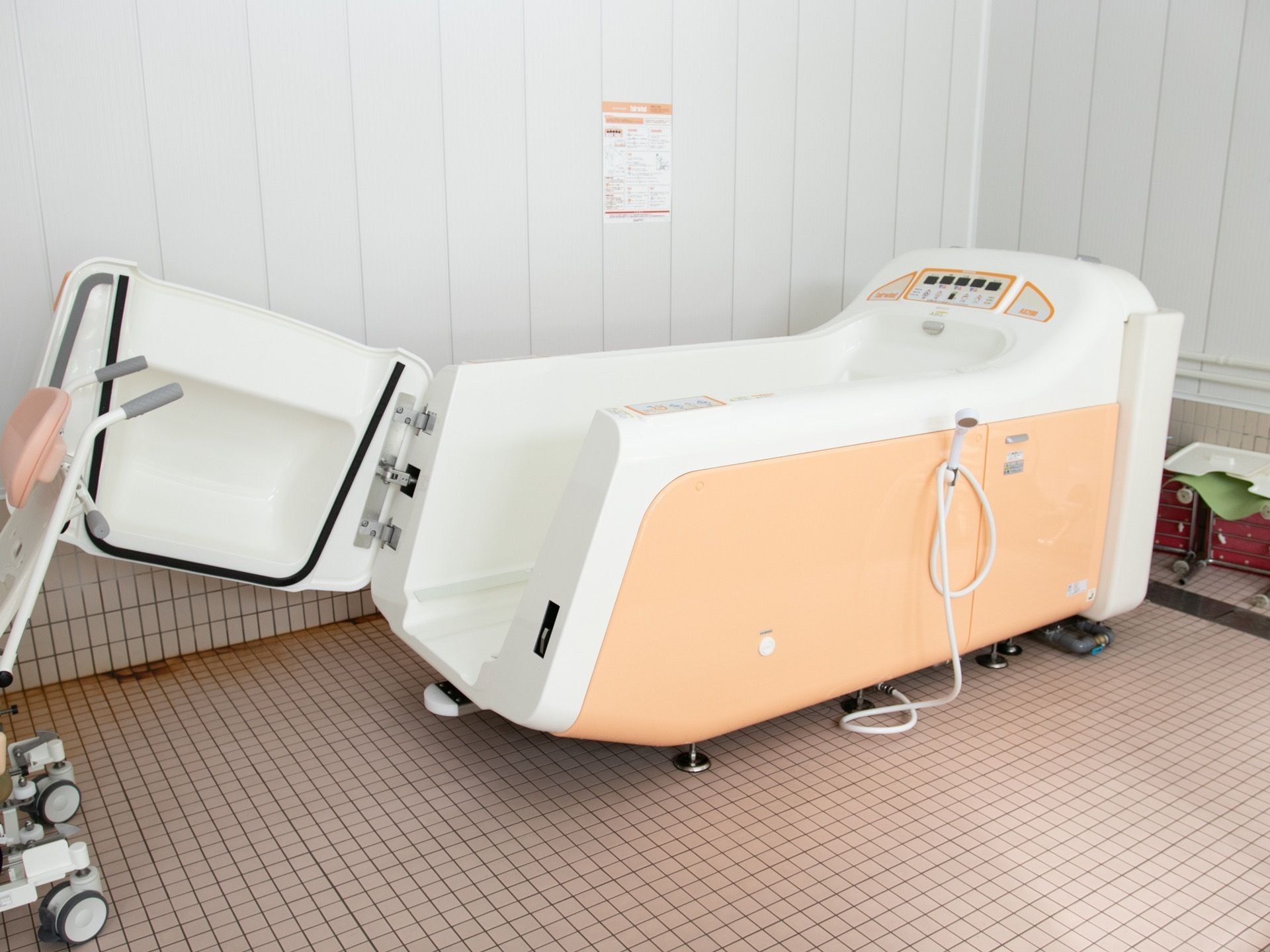 浴室内に置かれた白と淡いオレンジ色のボディの介護浴槽である。扉が開いた状態になっている。搬送車が待機している。