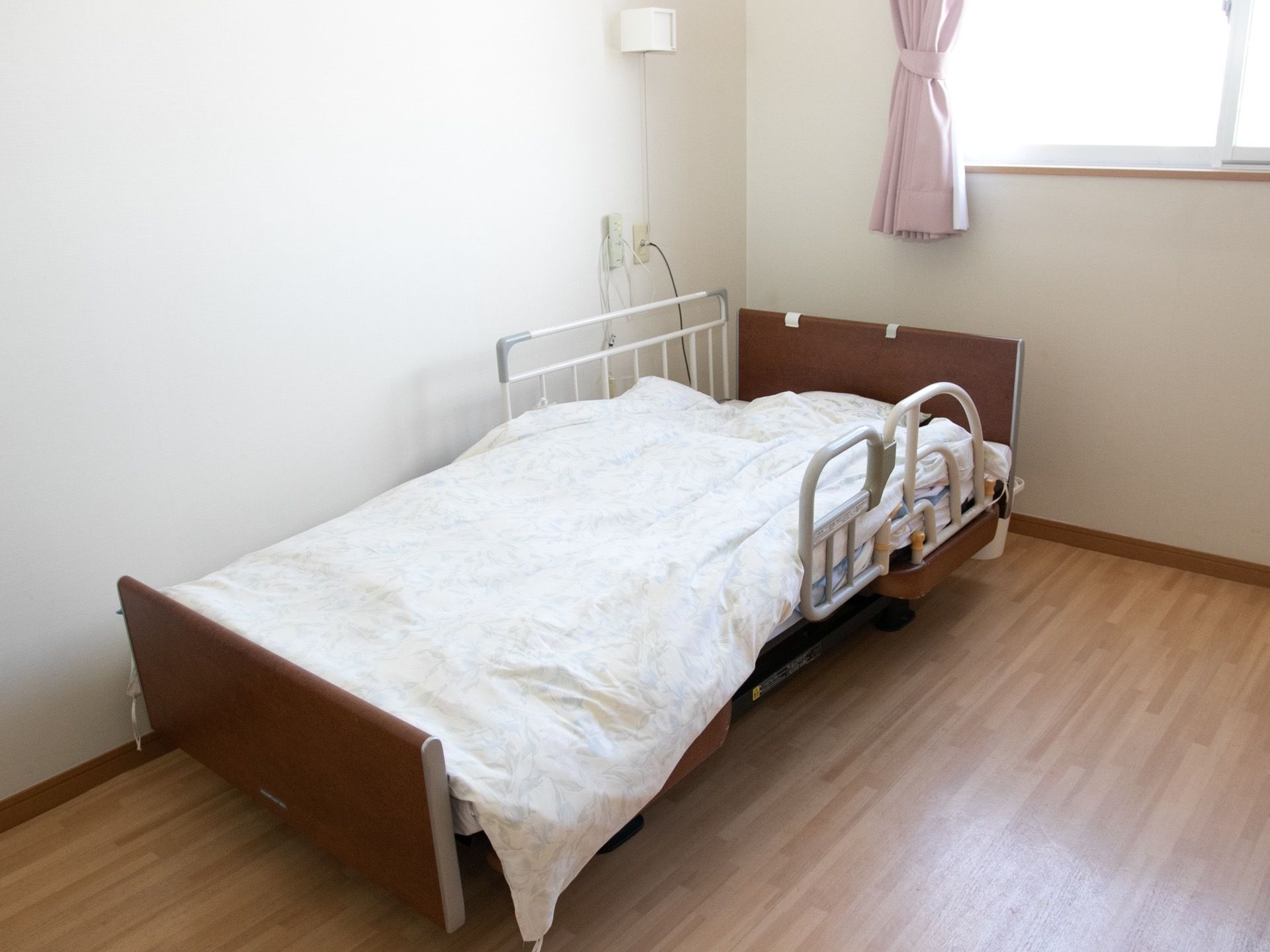 段差のないフローリング張りの居室内に、介護用のベッドが設置されている。ベッドガードが取り付けられ、カバーリングされた布団が敷かれている。