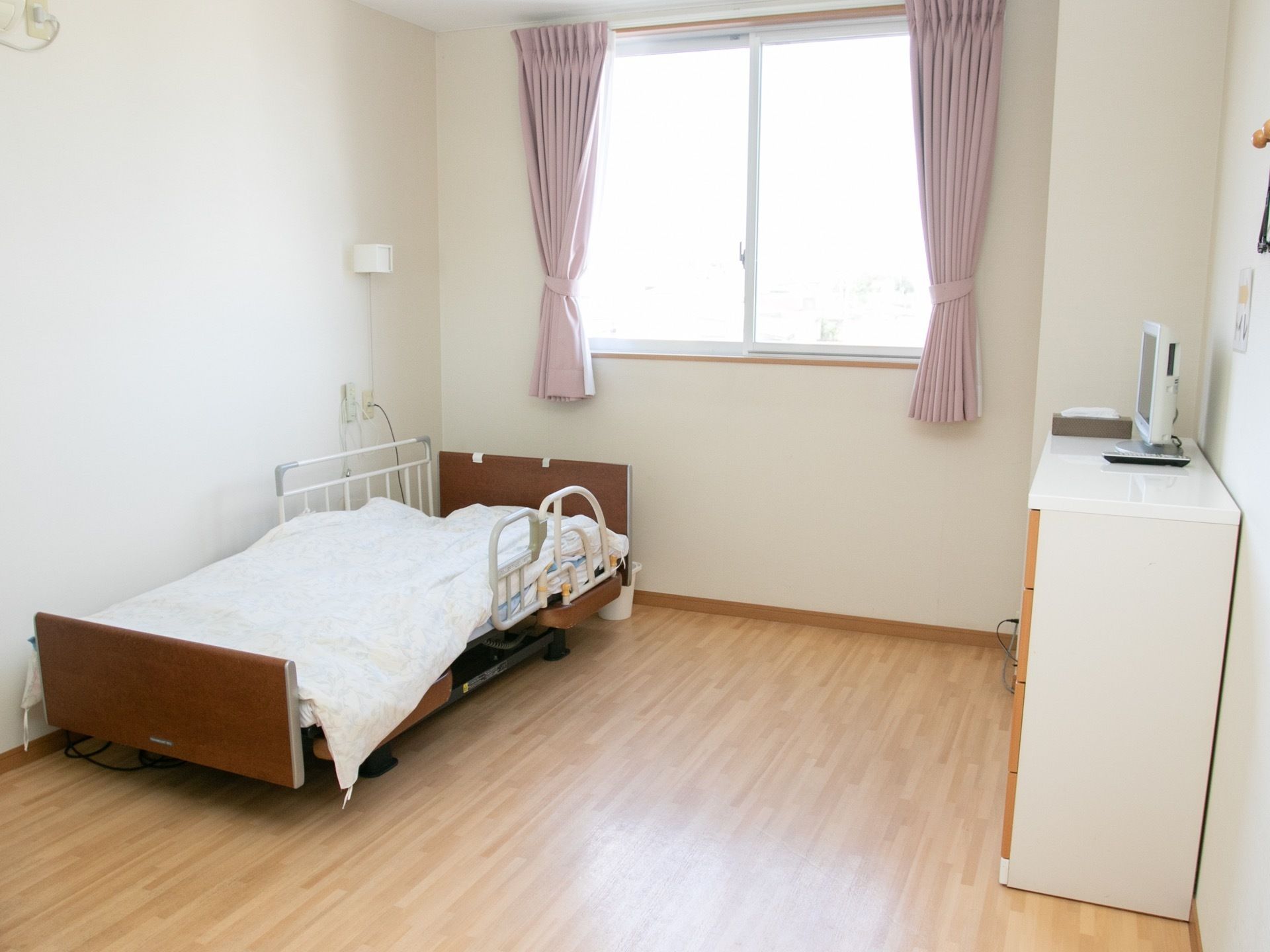 明るい色のフローリングの室内に、介護ベッドとタンスが置かれている。タンスの上には小さなテレビが載っている。