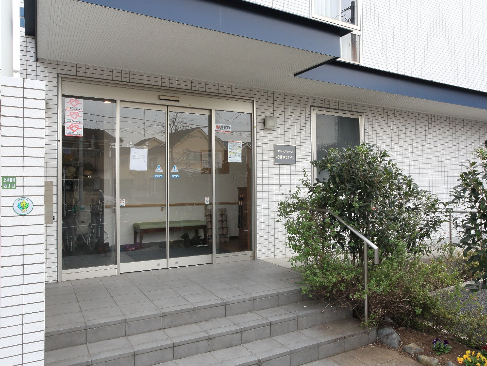 外から見た正面玄関口の様子である。ガラス張りのスライド式自動ドアになっており、AEDを備えている旨のステッカーが貼ってある。