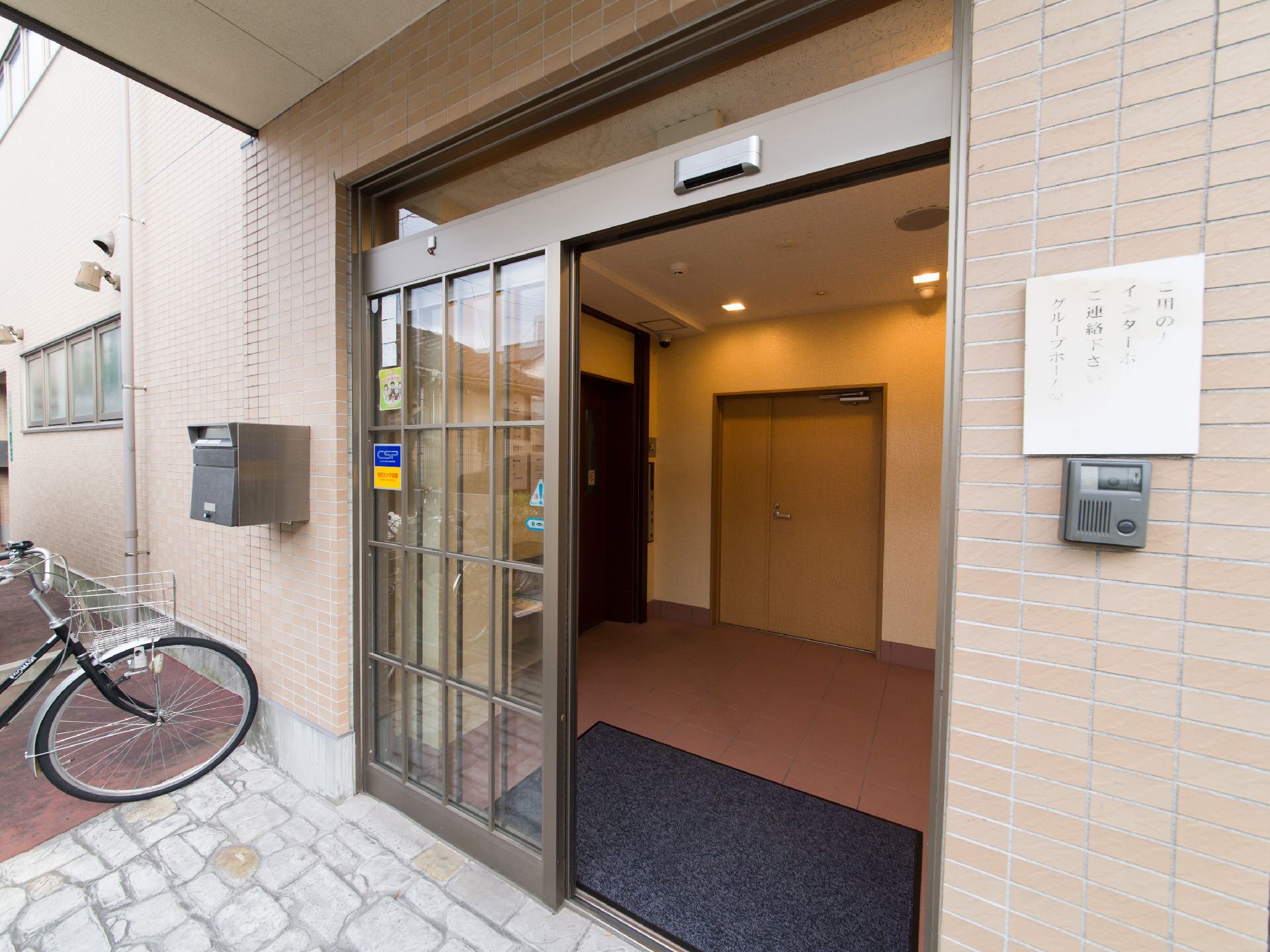 施設の玄関ドアはブラウンの格子にガラスがはめ込まれた自動ドアである。右側の壁にはインターホンがあり、上部には説明が書かれている。